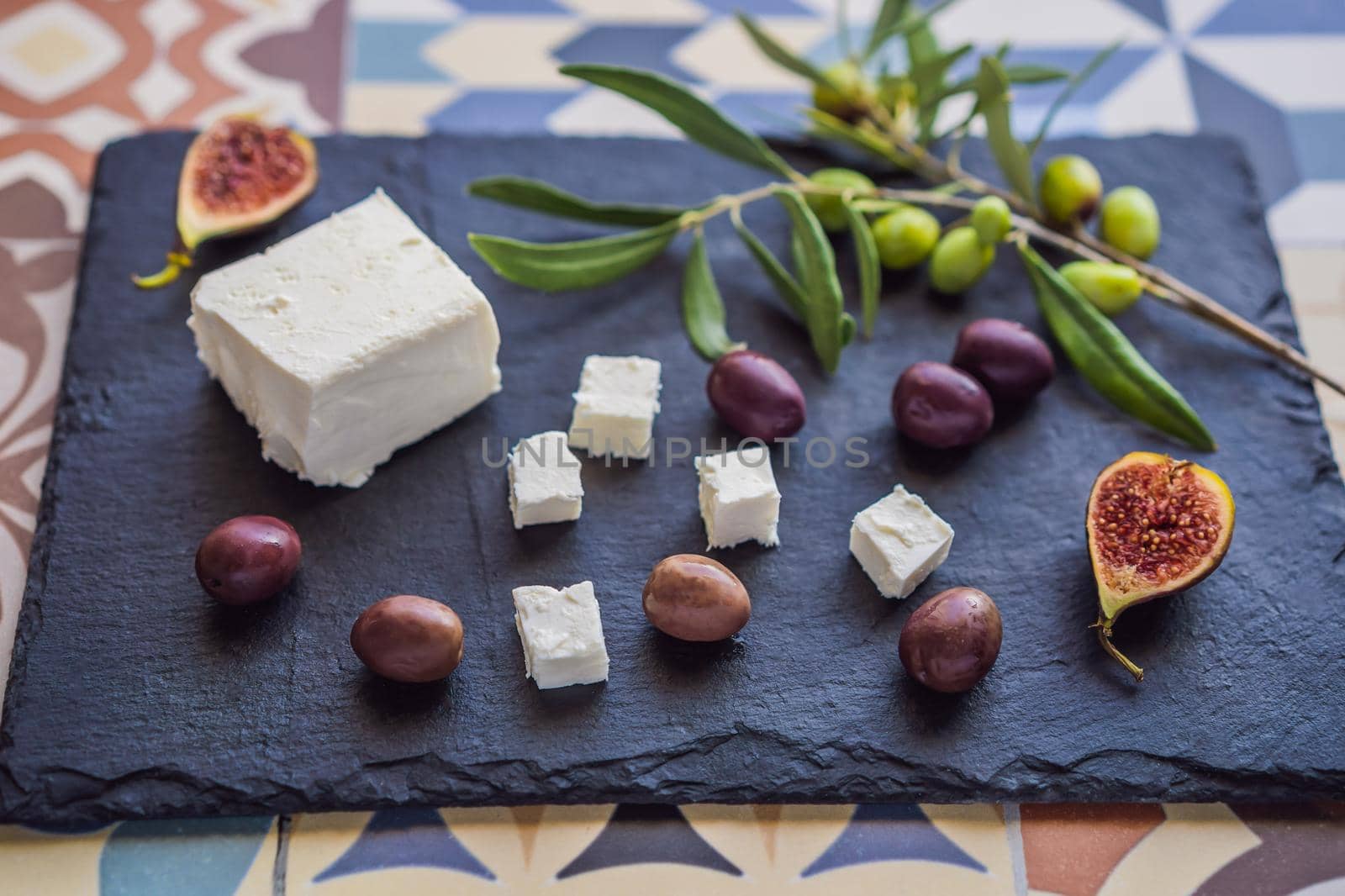 Green and black olives with loaf of fresh bread, feta cheese and young olives branch on olive wood chopping board over dark background.