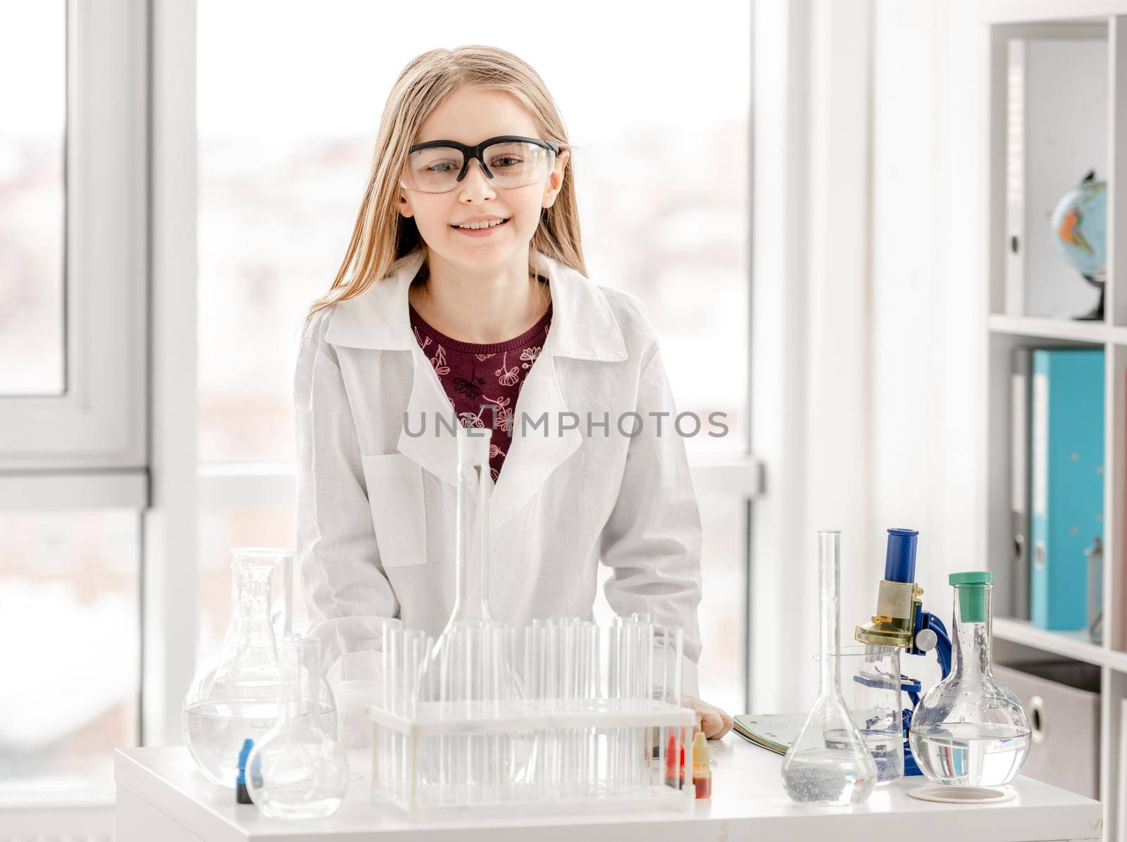Smart girl during scientific chemistry experiment wearing protection glasses. Schoolgirl with equipment and chemical liquids looking at camera
