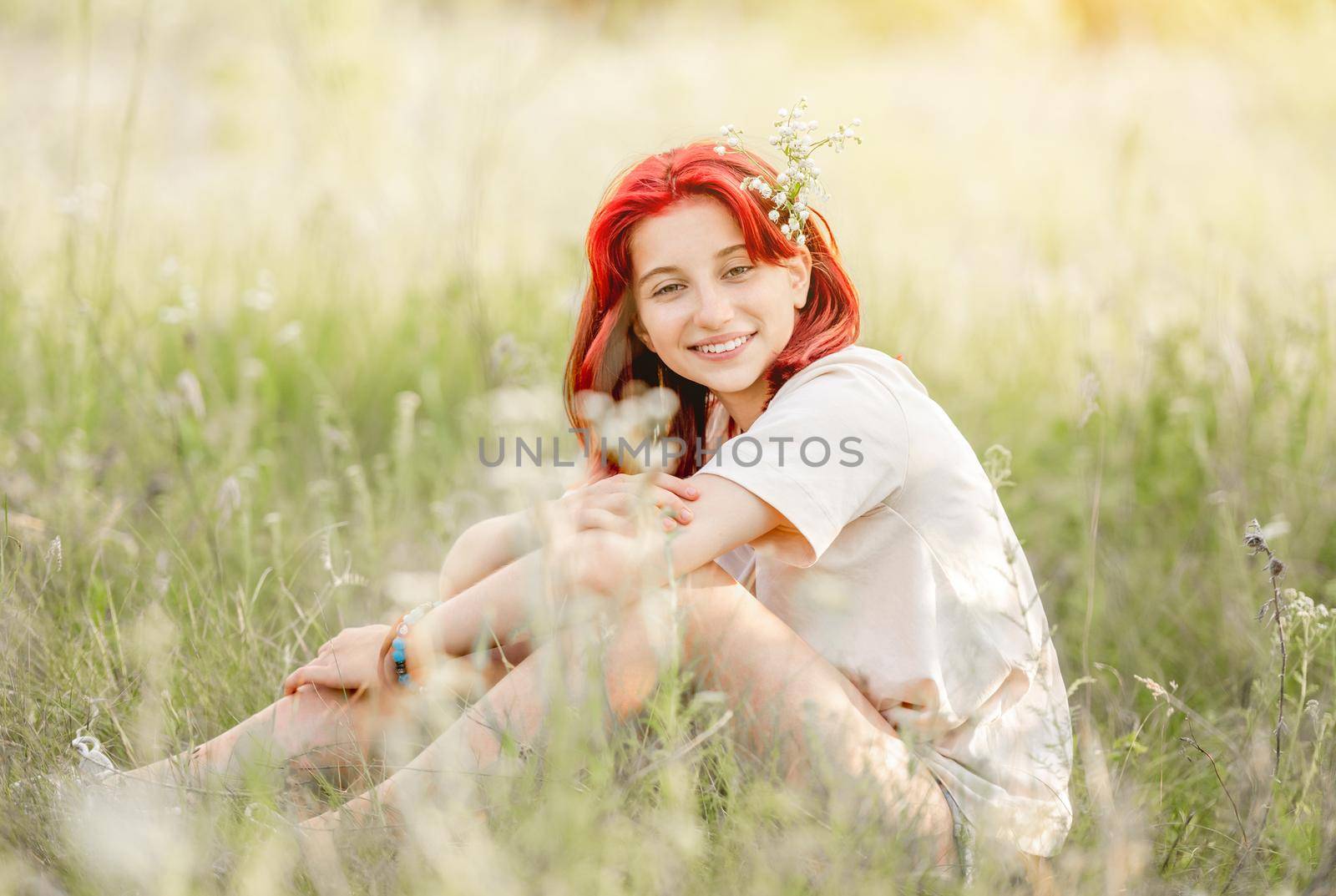Girl with red hair sitting at the nature and smiling. Pretty young female in the sunny field with flowers