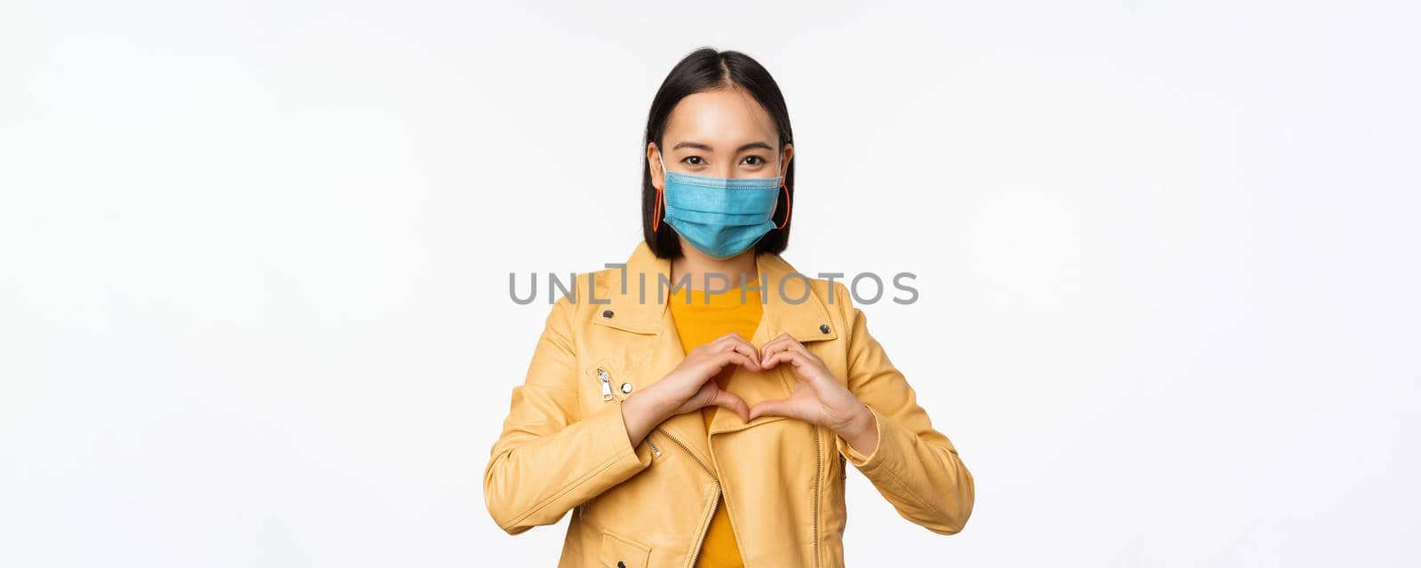 Covid-19 and people concept. Young asian woman in medical face mask, showing heart gesture and smiling with care, white background.