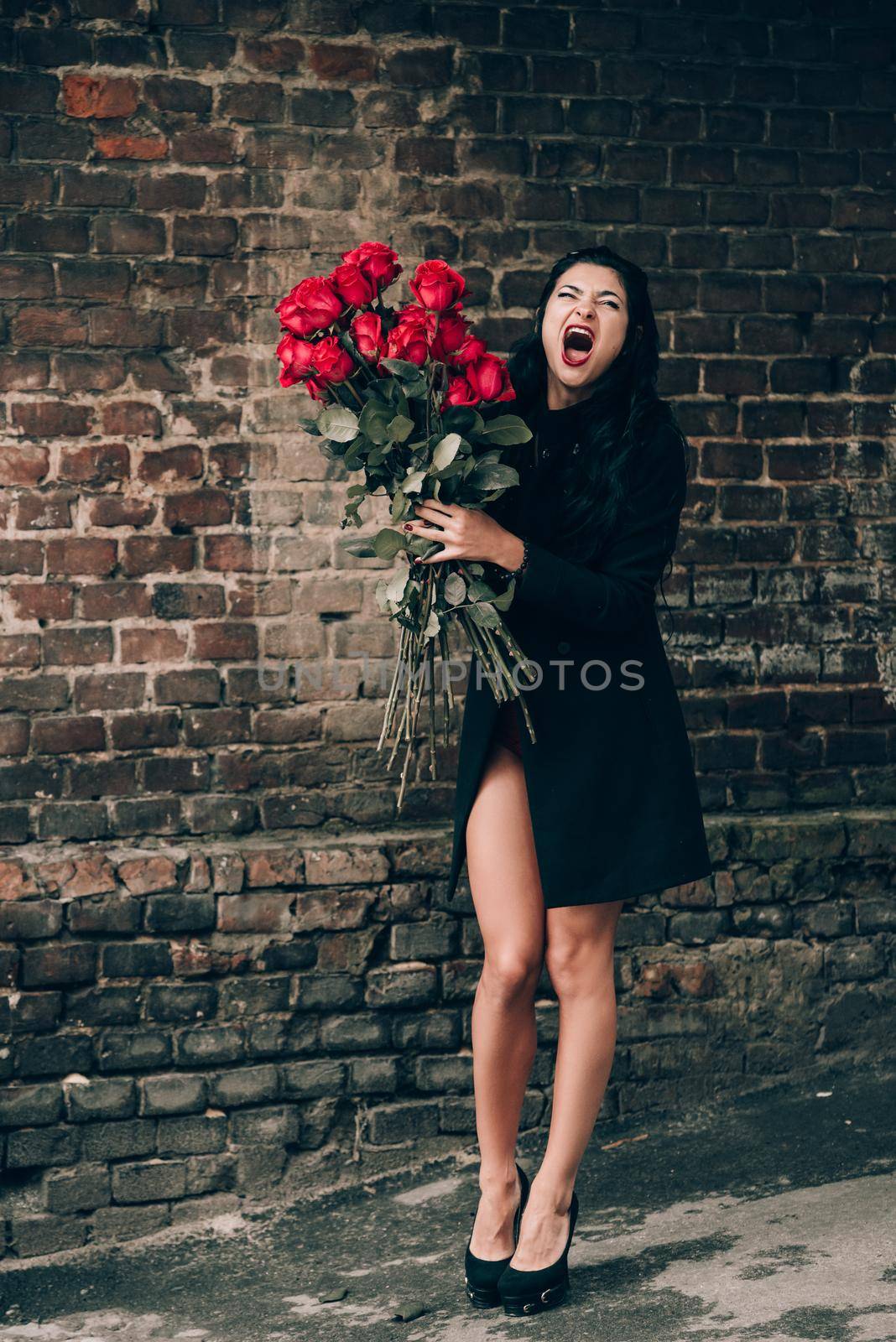 Elegant beautiful brunette woman in red shorts and black coat posing near brick wall with a big bouquet of red roses. Birthday. March 8. Valentines day.