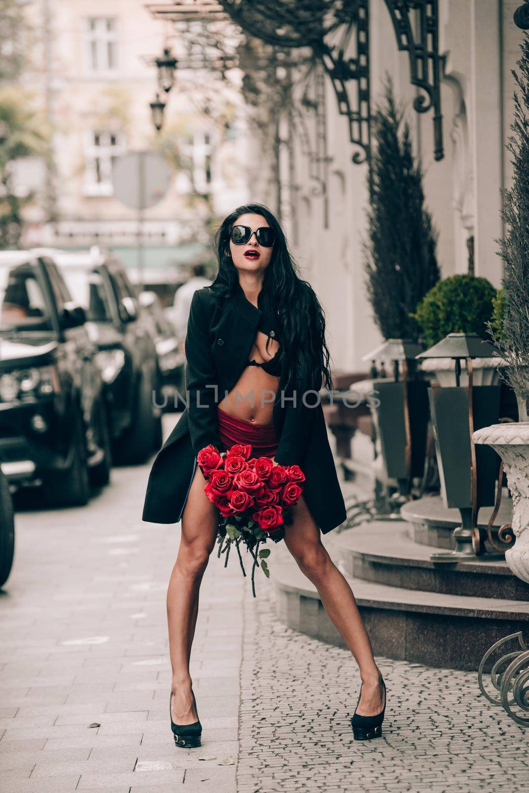 Elegant beautiful brunette woman in red shorts and black coat posing on a city streets with a big bouquet of red roses. Birthday. Valentines day.