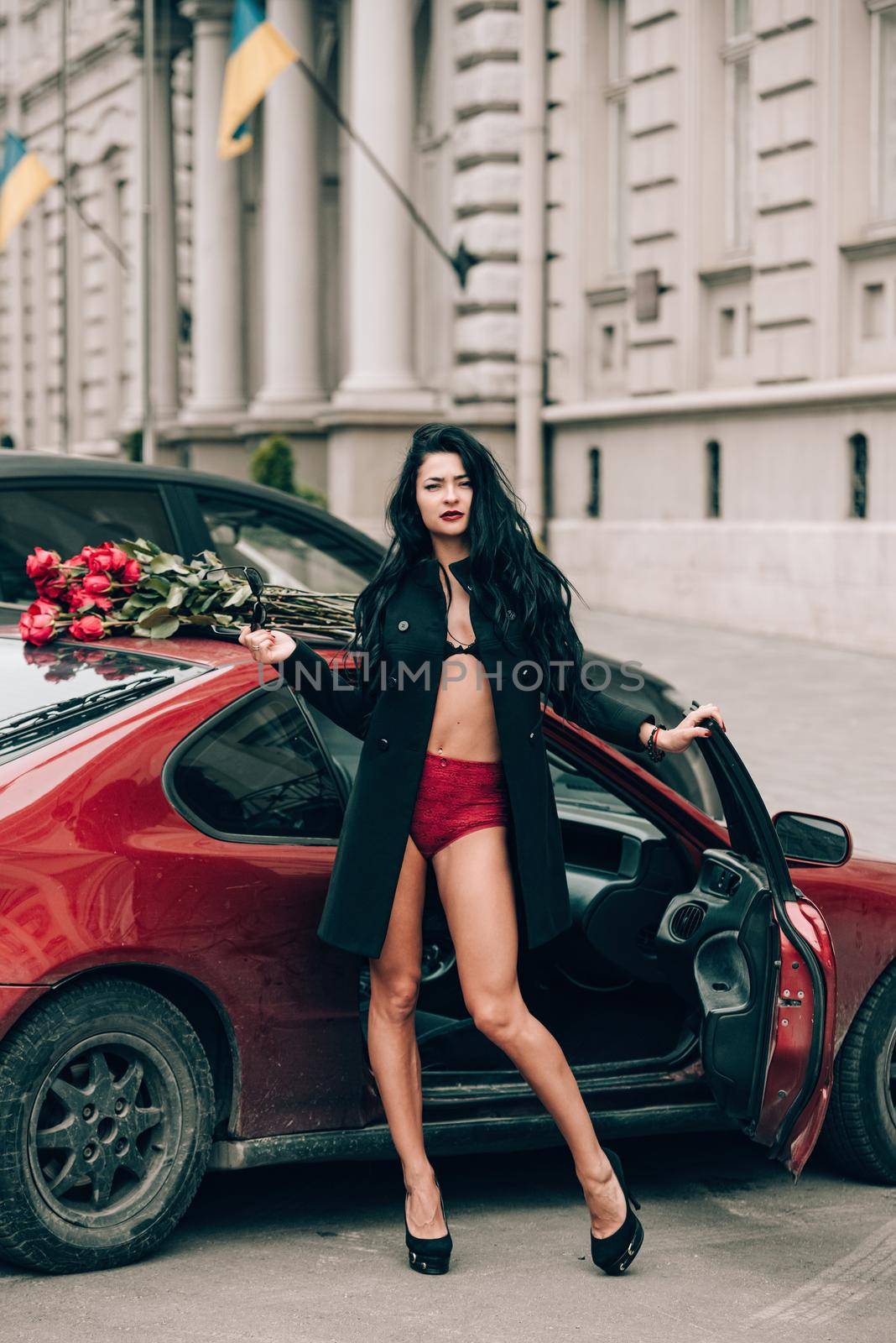 Elegant beautiful brunette woman in red shorts and black coat posing near red car. big bouquet of red roses on a car roof. Birthday. March 8.