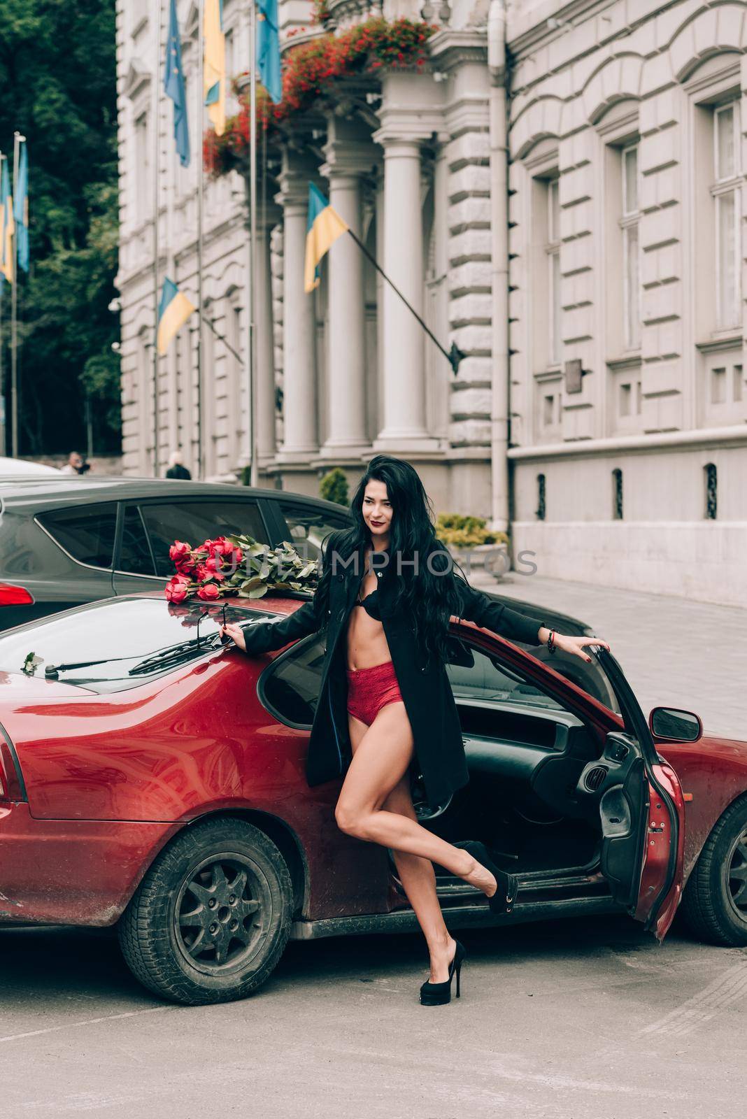 Elegant beautiful brunette woman in red shorts and black coat posing near red car. big bouquet of red roses on a car roof. Birthday. March 8.