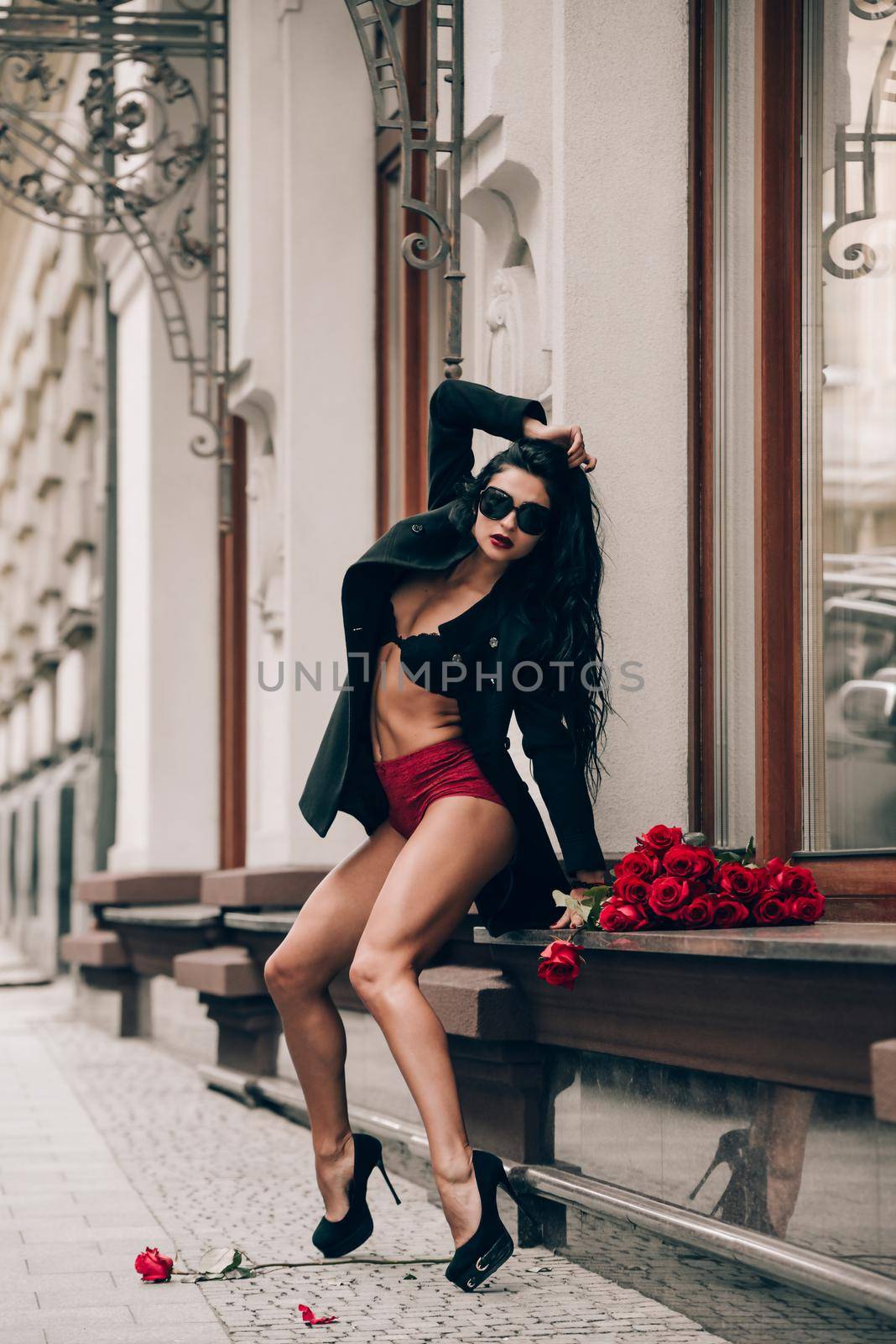 Elegant beautiful brunette woman in sunglasses and red shorts and black coat girl posing sitting on the windowsill with a big bouquet of red roses. by Ashtray25