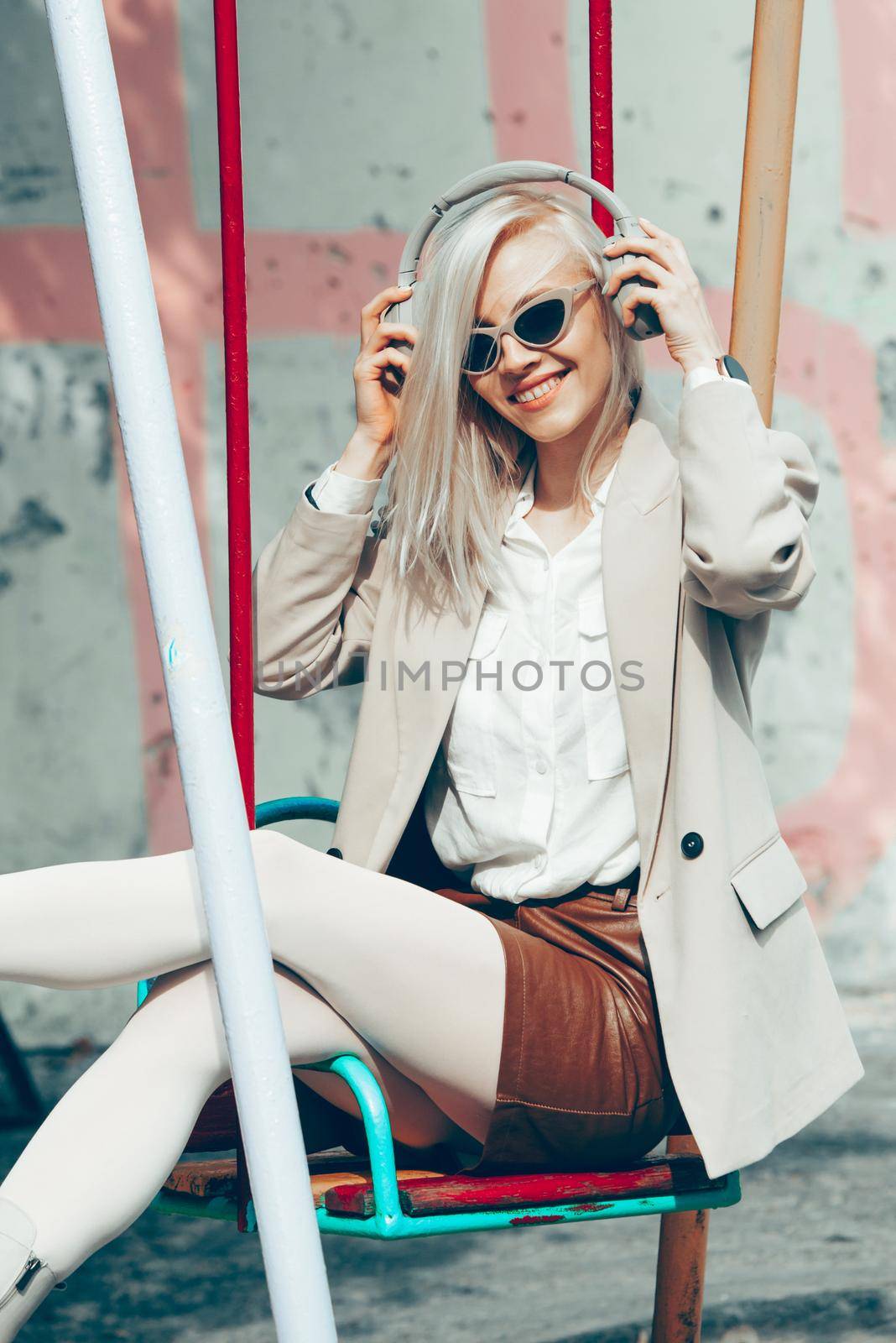 Portrait of blonde hipster fashion model wear stylish wireless headphones enjoy listen new cool music. woman wearing white jacket, blouse, tights and brown leather skirt