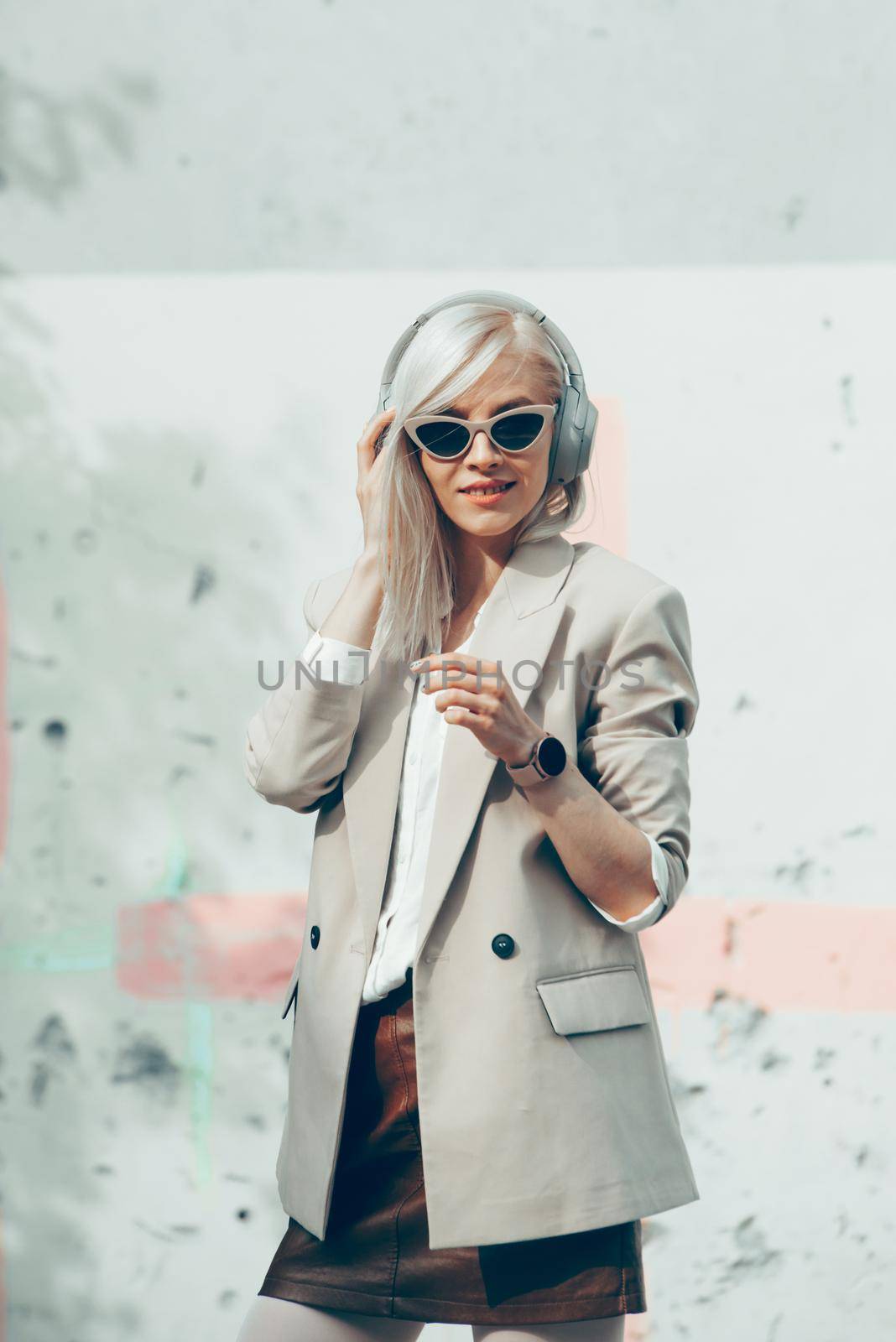 Portrait of blonde hipster fashion model wear stylish wireless headphones enjoy listen new cool music. woman wearing white jacket, blouse, tights and brown leather skirt