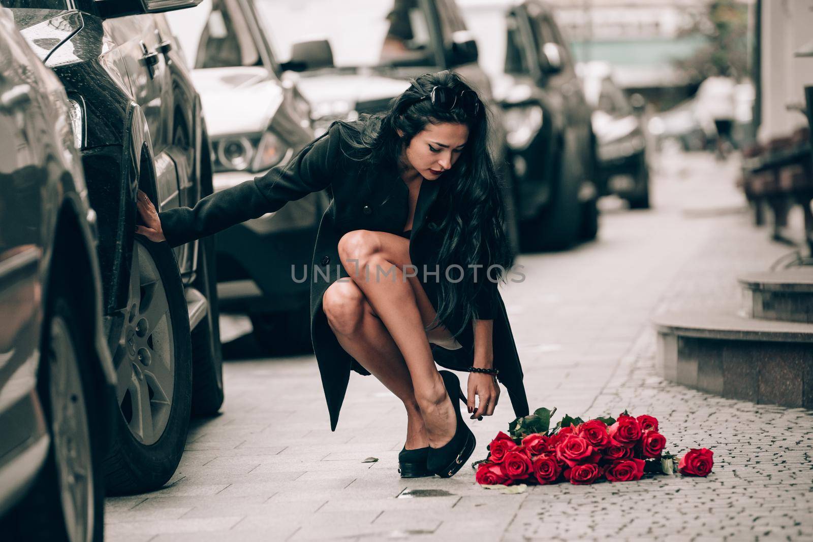 Elegant beautiful brunette woman in red shorts and black coat posing on a city streets with a big bouquet of red roses. Birthday. Valentines day.