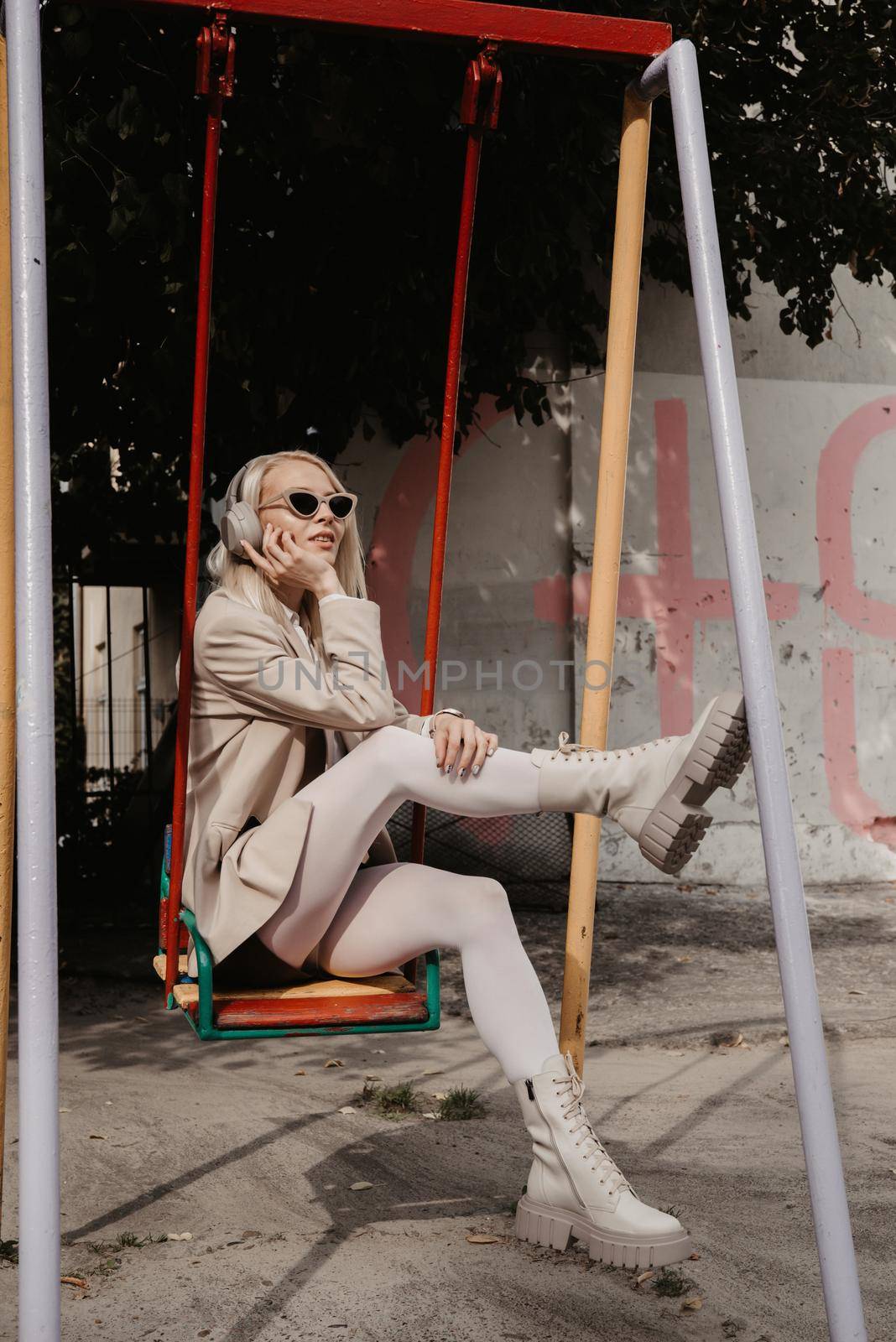 Portrait of blonde hipster fashion model wear stylish wireless headphones enjoy listen new cool music. woman wearing white jacket, blouse, tights and brown leather skirt