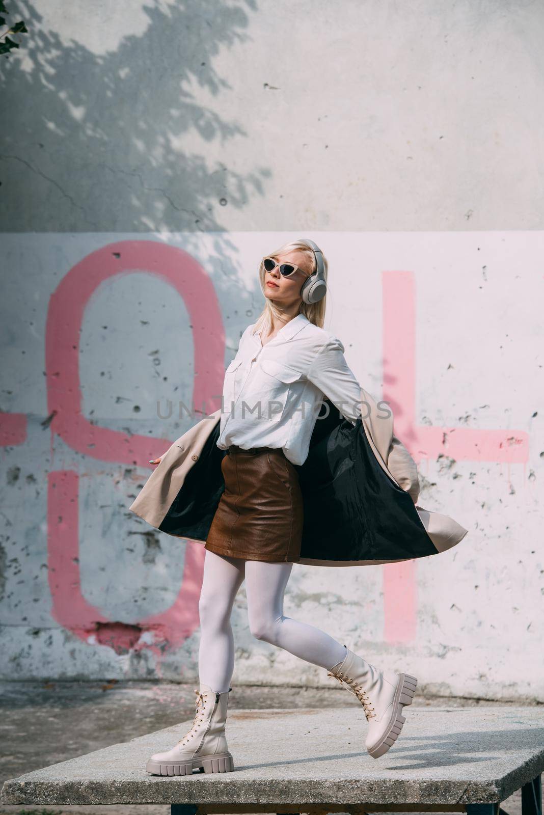 Portrait of blonde woman in headphones and sunglasses listening music. woman wearing white jacket, blouse, tights and brown leather skirt. white high leather boots