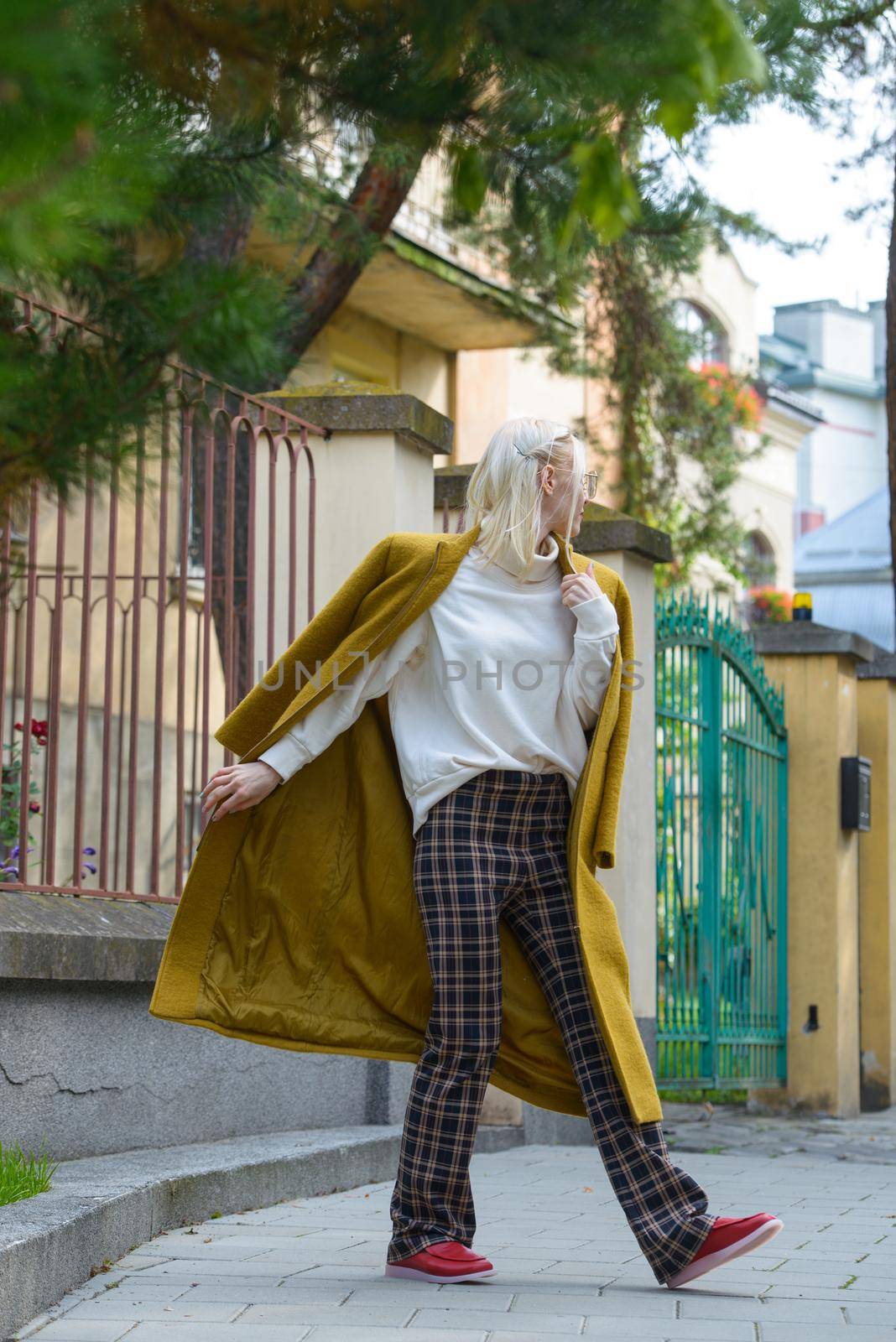 Fashionable beautiful young woman with blond hair in a stylish long coat, checkered pants, red shoes and glasses poses in the city streets. Feminine urban style.