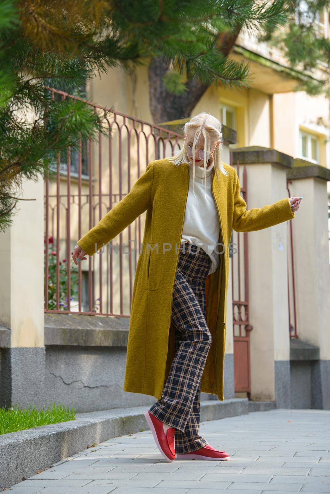 Fashionable beautiful young woman with blond hair in a stylish long coat, checkered pants, red shoes and glasses poses in the city streets. Feminine urban style.