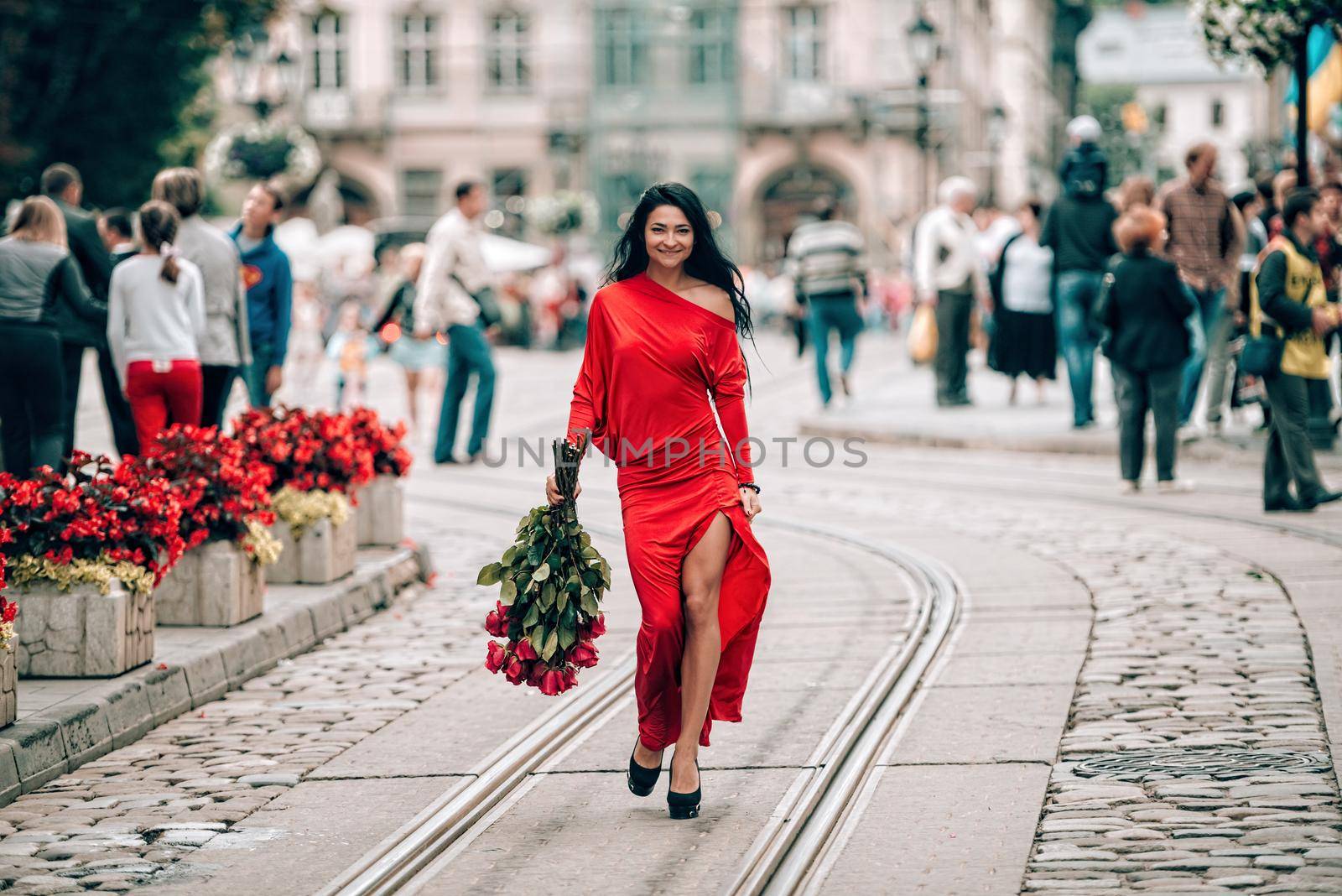 Charming young woman in red sexy dress posing with a bouquet of red roses. photo of a seductive woman with black hair on the city streets. Selective focus, filmgrain.