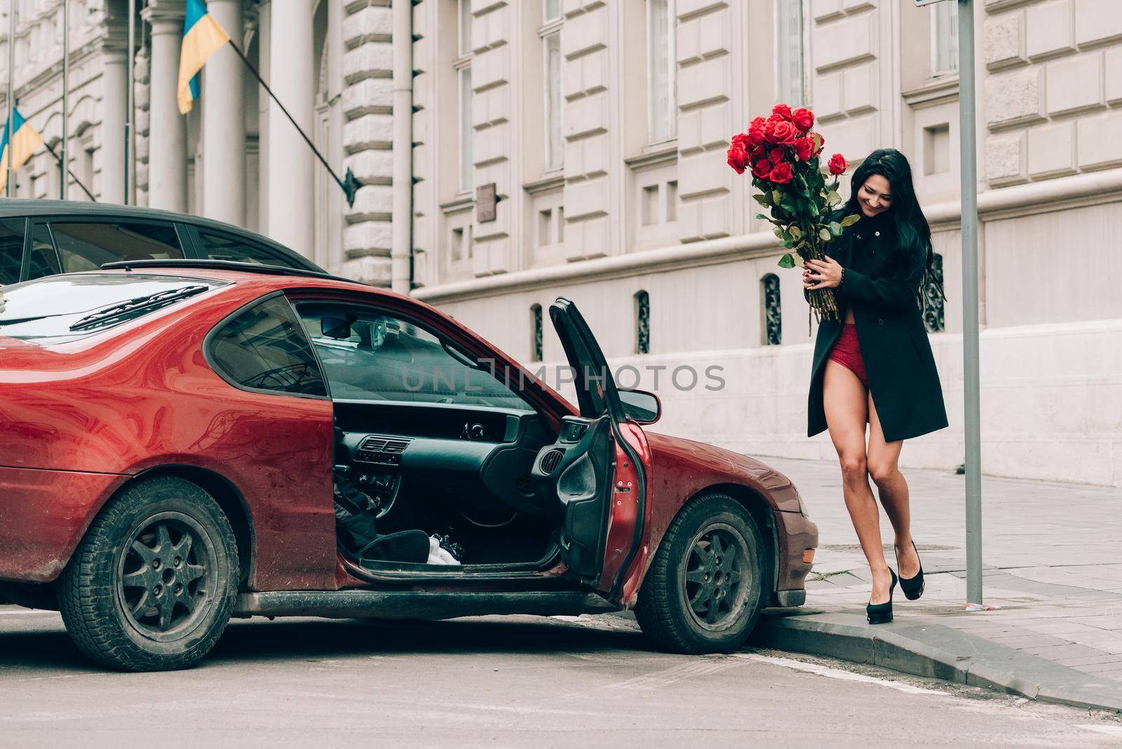 Elegant beautiful brunette woman in red shorts and black coat posing near red car. big bouquet of red roses. Birthday. March 8. Valentines day. by Ashtray25