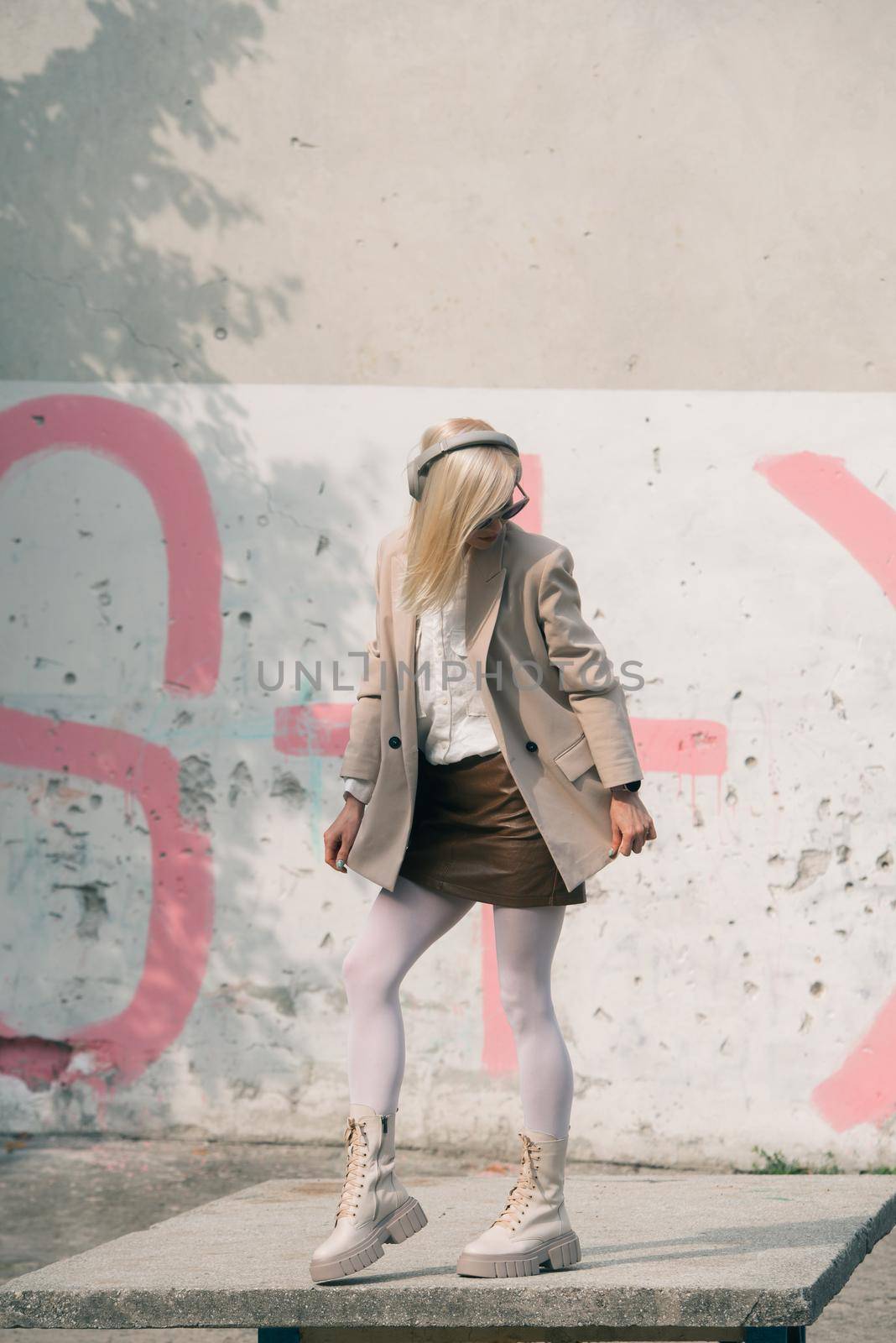 Portrait of blonde woman in headphones and sunglasses listening music. woman wearing white jacket, blouse, tights and brown leather skirt. white high leather boots