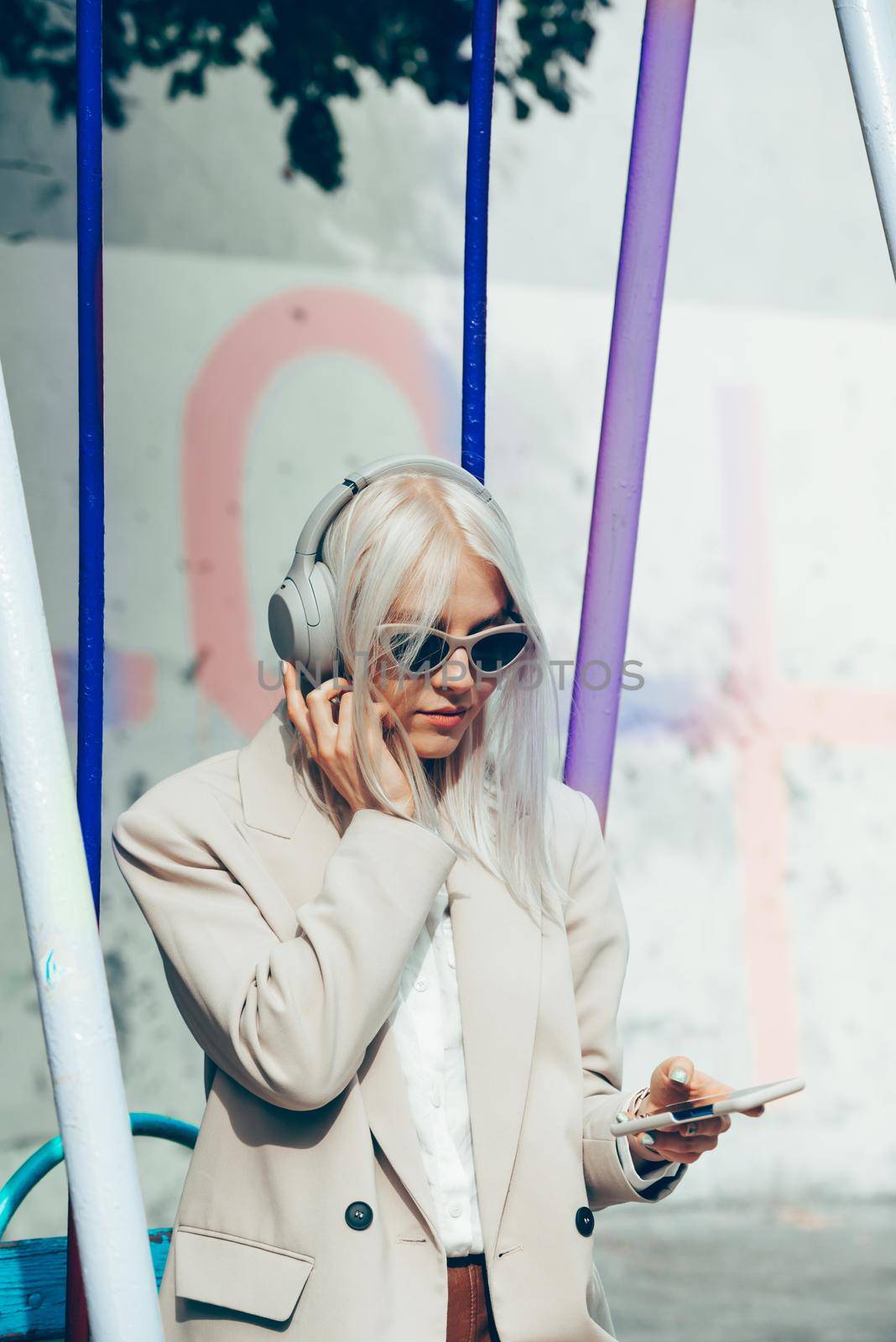 Portrait of blonde hipster fashion model wear stylish wireless headphones enjoy listen new cool music. woman wearing white jacket, blouse, tights and brown leather skirt