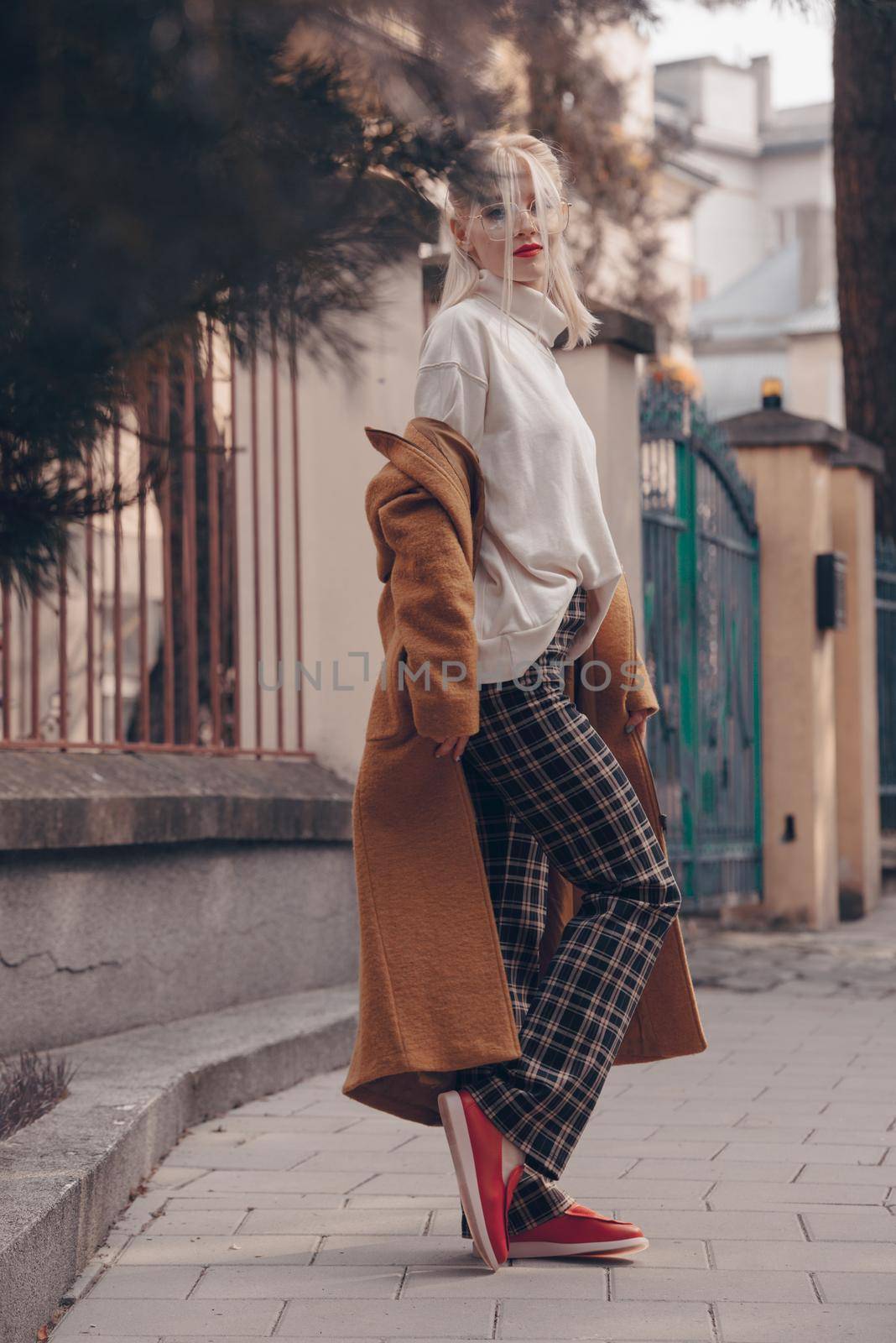 Fashionable beautiful young woman with blond hair in a stylish long coat, checkered pants, red shoes and glasses poses in the city streets. Feminine urban style.