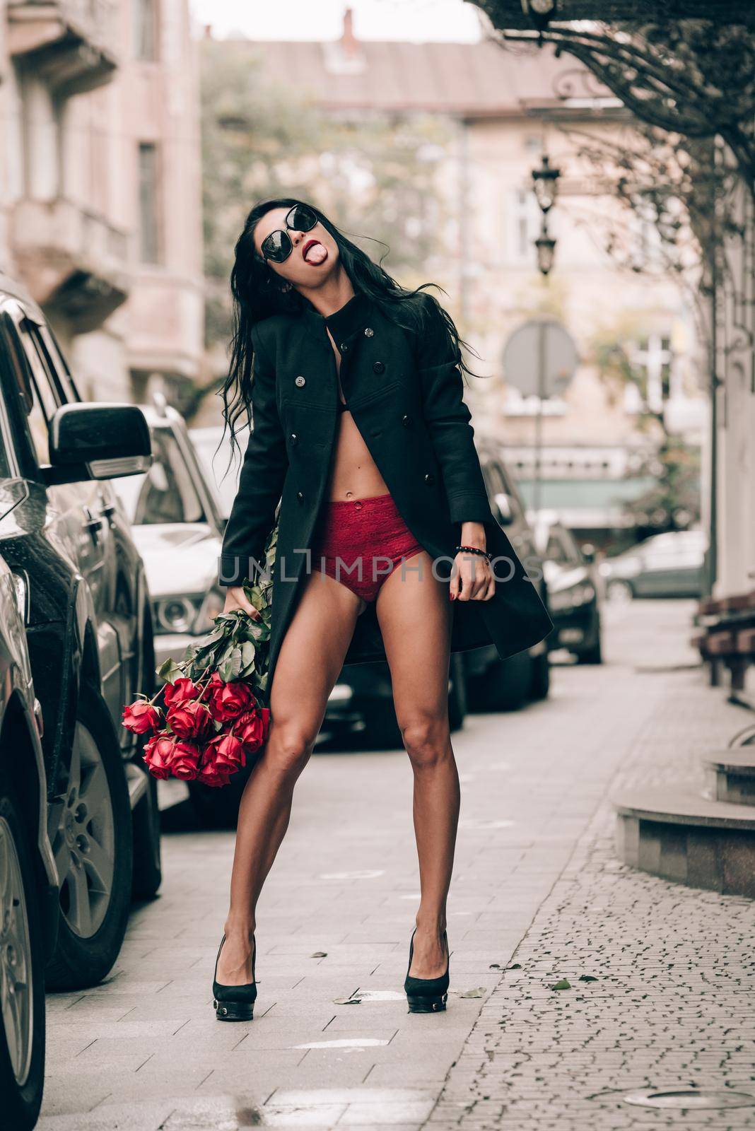 Elegant beautiful brunette woman in sunglasses and red shorts and black coat posing on a city streets with a big bouquet of red roses. by Ashtray25