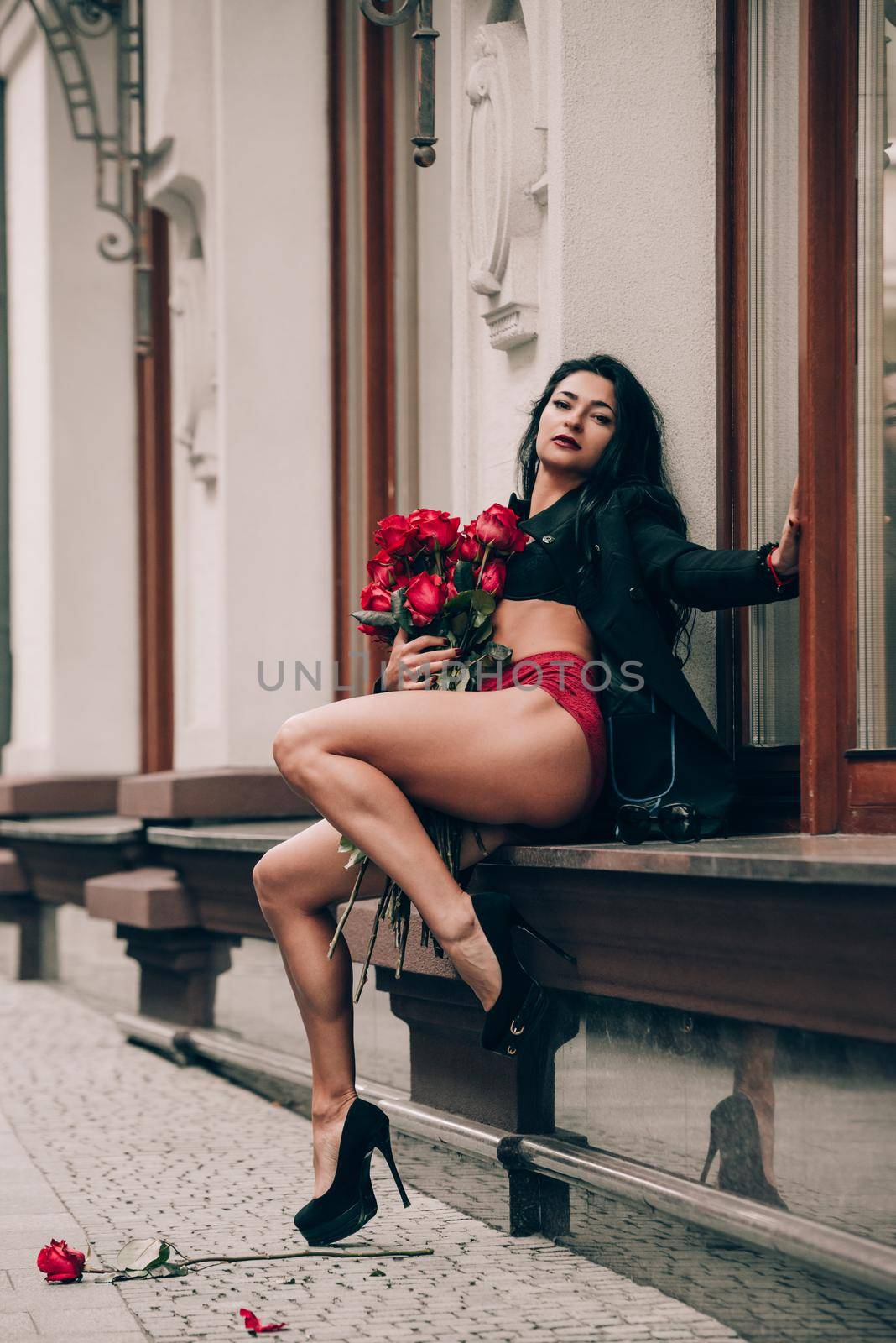 Elegant beautiful brunette woman in red shorts and black coat girl posing sitting on the windowsill with a big bouquet of red roses. Birthday. by Ashtray25