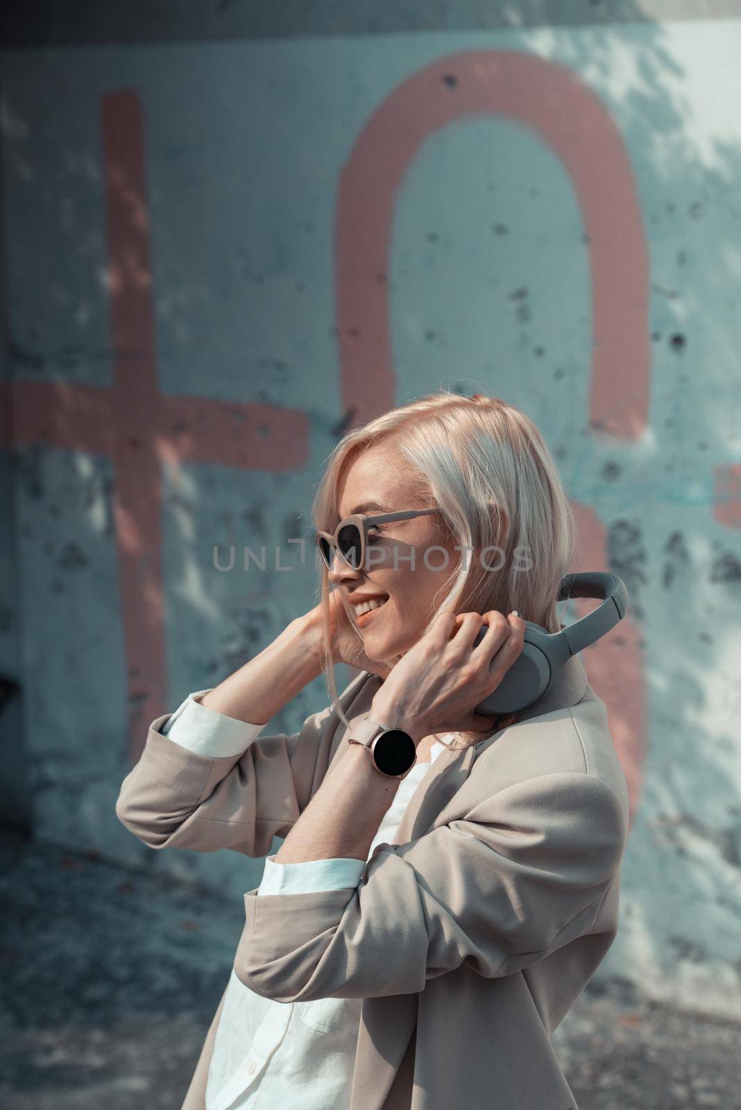 Portrait of blonde woman with headphones in hands. . woman wearing white jacket, blouse, tights and brown leather skirt