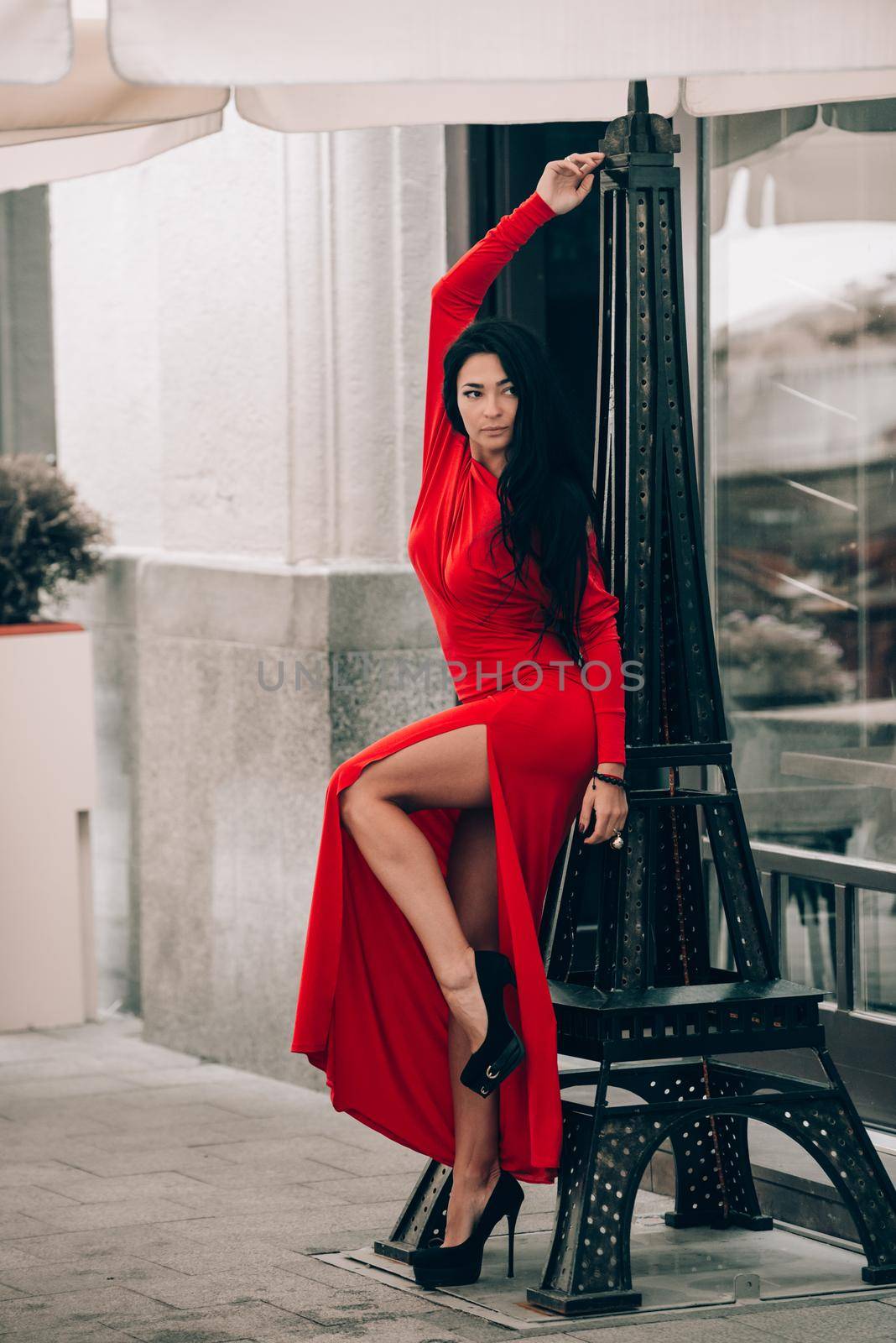 Charming young woman in red sexy dress posing . photo of a seductive woman with black hair near decorative tower. Selective focus
