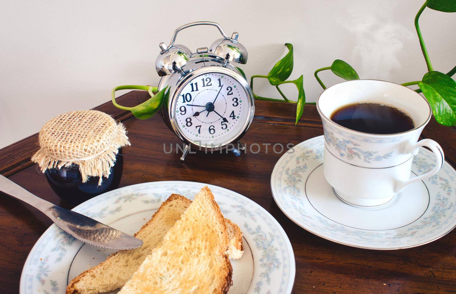 Breakfast table with tea or coffee in a dainty cup and saucer with slices of toast and a pot of jam and a retro alarm clock