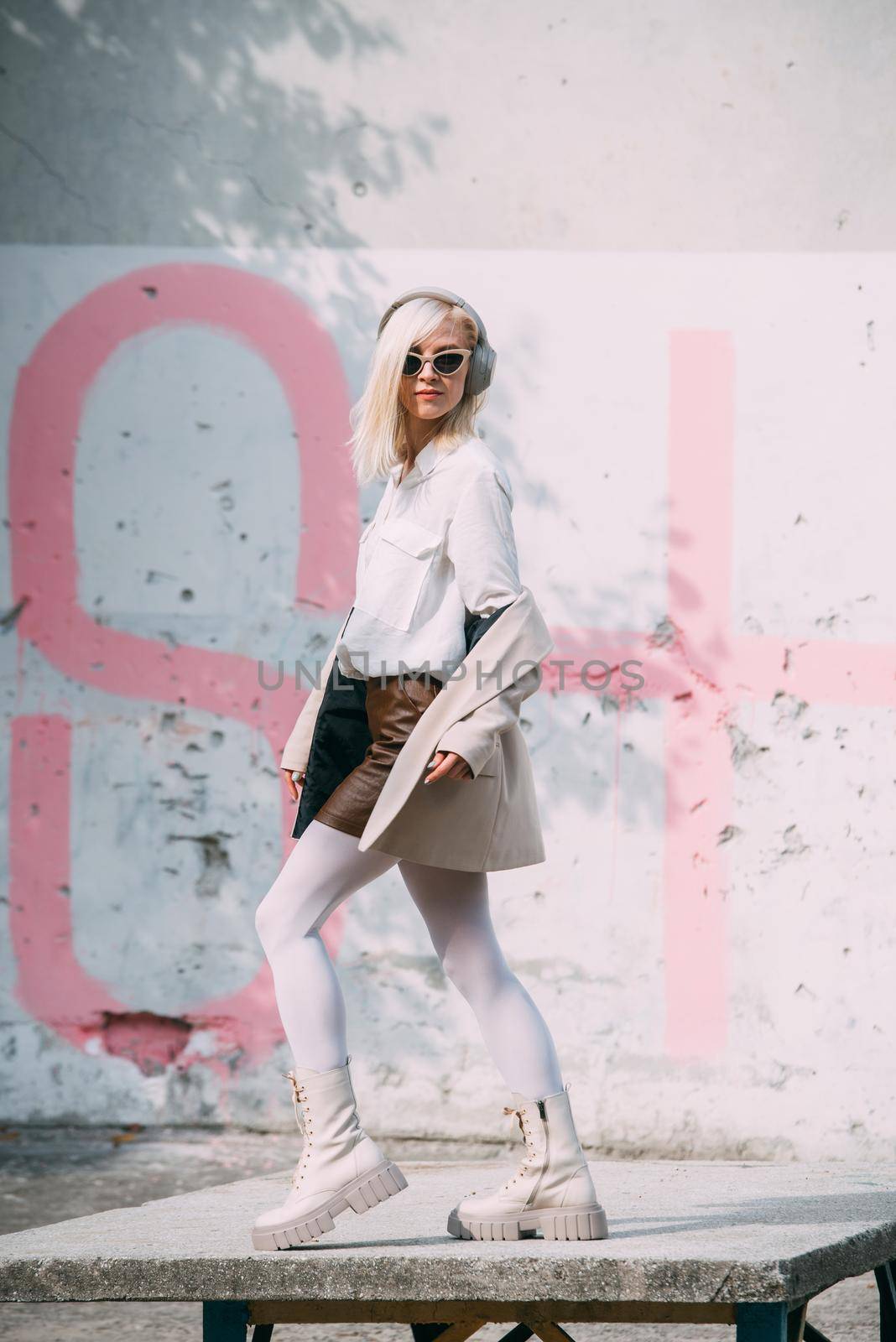Portrait of blonde woman in headphones and sunglasses listening music. woman wearing white jacket, blouse, tights and brown leather skirt. white high leather boots