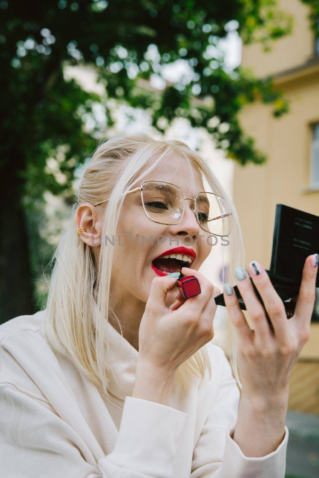 young smiling hipster happy woman in glasses doing make-up using lipstick and mirror outdoors