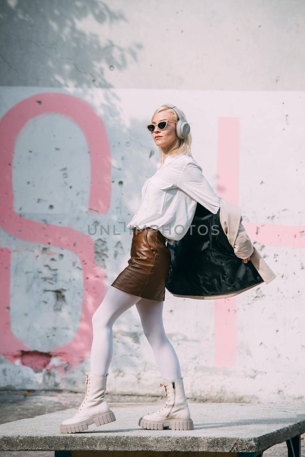 Portrait of blonde woman in headphones and sunglasses listening music. woman wearing white jacket, blouse, tights and brown leather skirt. white high leather boots