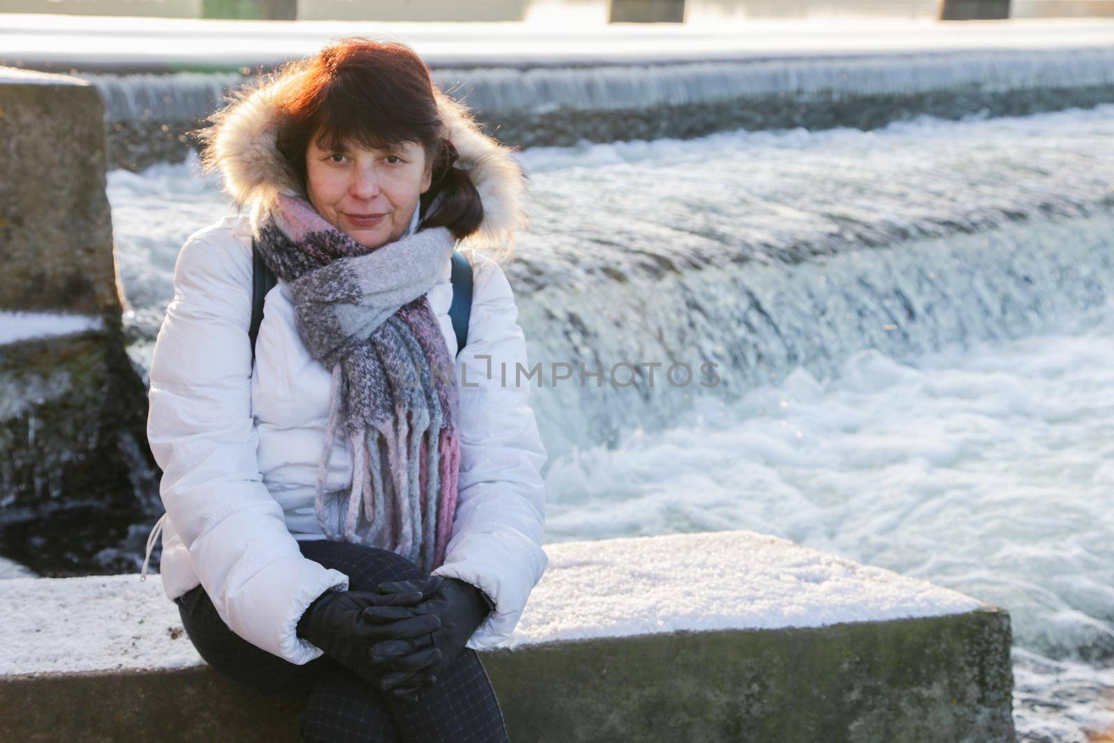 Belt portrait of a sitting pretty older woman in a white jacket with a scarf and black gloves looking at the camera.