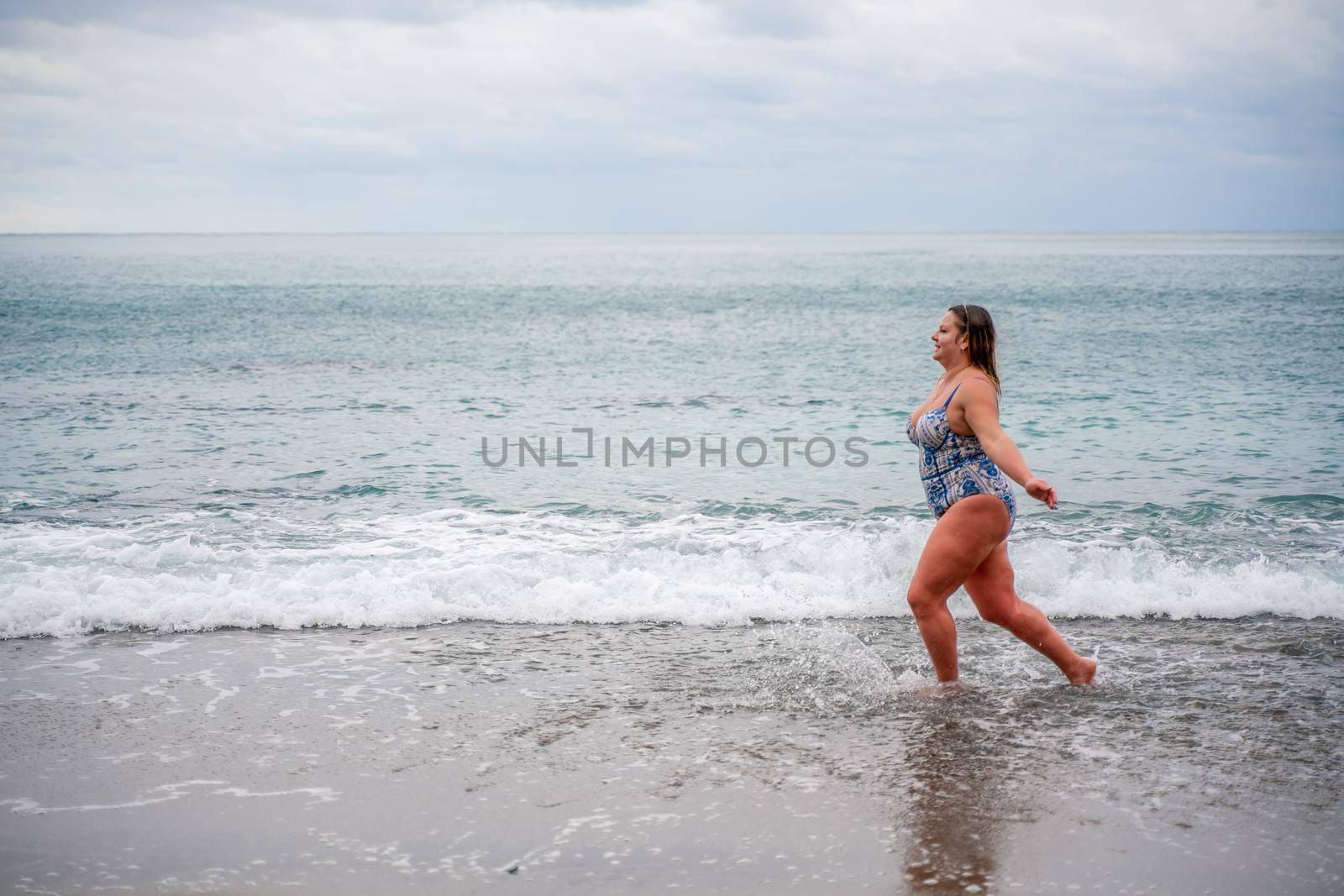 A plump woman in a bathing suit enters the water during the surf. Alone on the beach, Gray sky in the clouds, swimming in winter