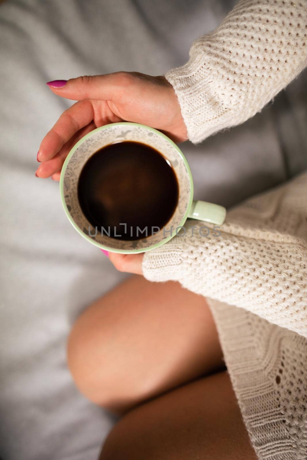 Close-up frame. A woman with slim legs sits on a bed in a living room and next to her holds a porcelain cup with hot coffee.