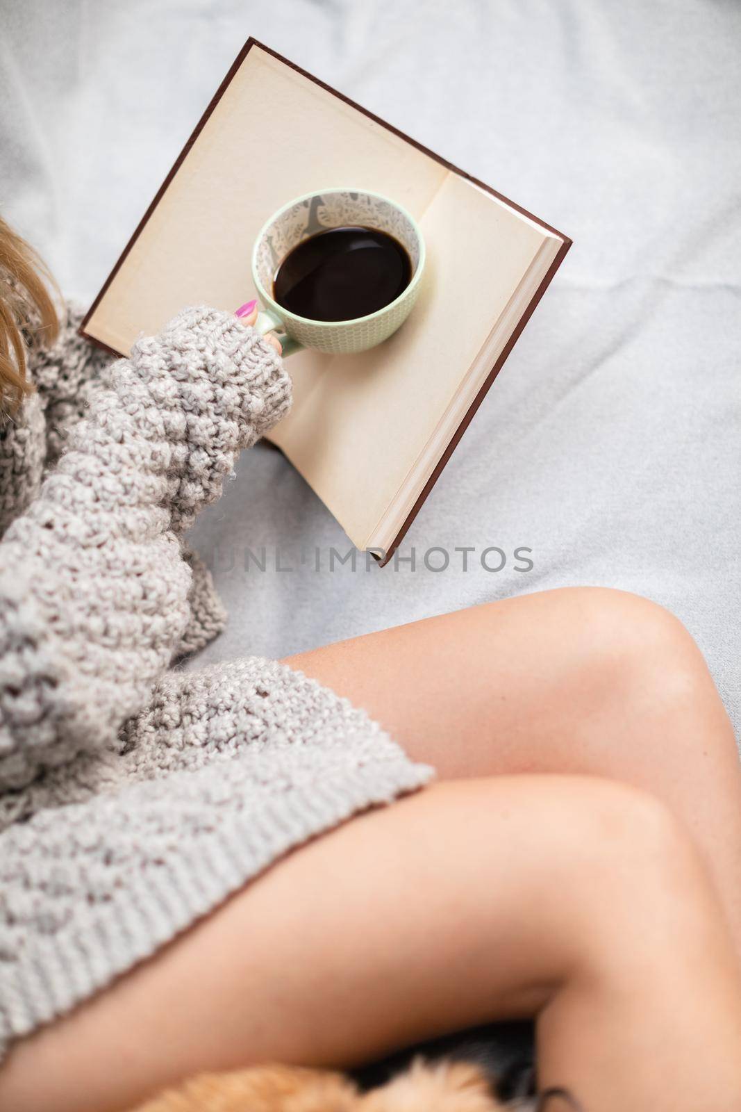 A woman lies on a bed with a book and a cup of coffee. Neat female legs with light complexion of skin. Aromatic coffee time while relaxing.
