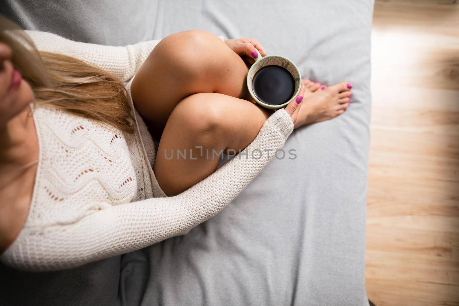 Cup of morning coffee held by female hand. Sexy female legs with light skin complexion. Relaxation in the comfort of your home.