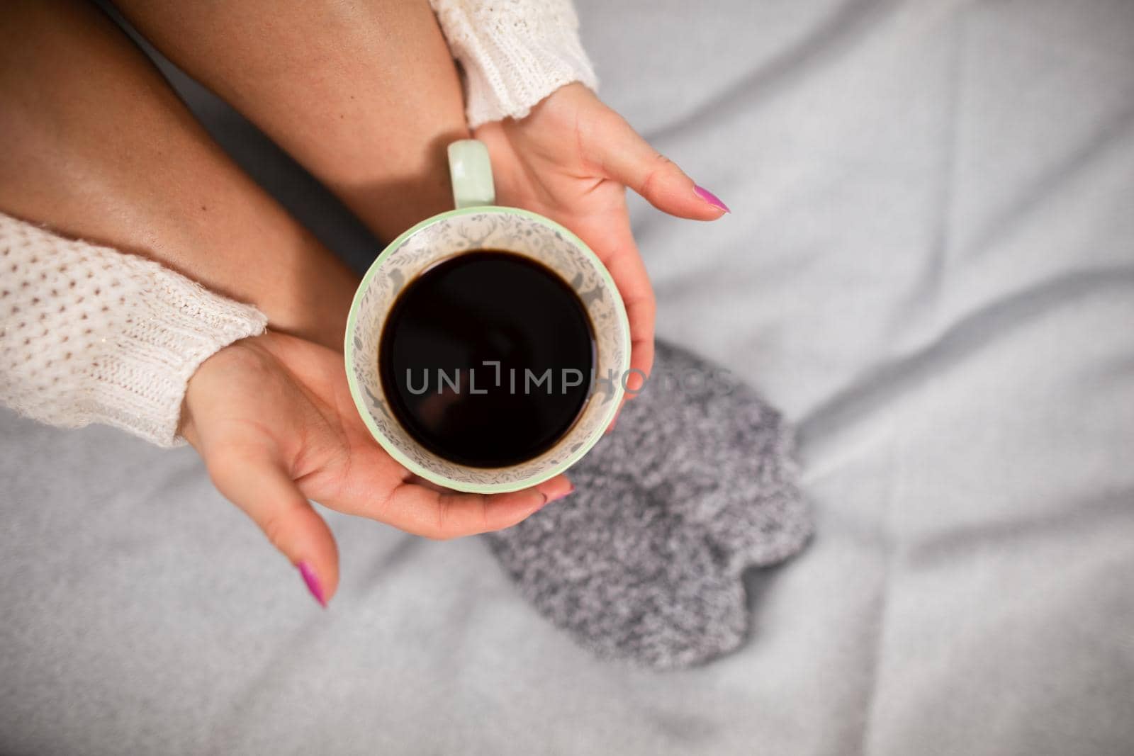 Porcelain cup with coffee held by woman's hands. Neat female legs. Relaxation in the comfort of home.