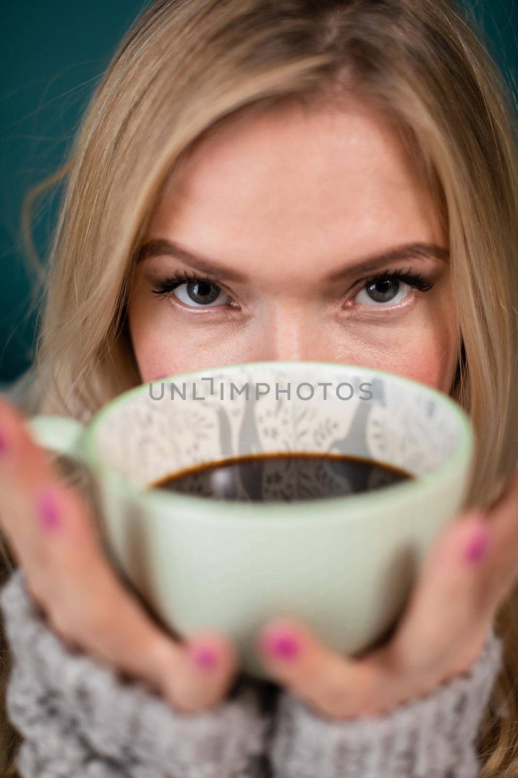 A large cup with hot coffee covers the face of a pretty young woman. Blonde woman during her morning ritual of drinking aromatic coffee.