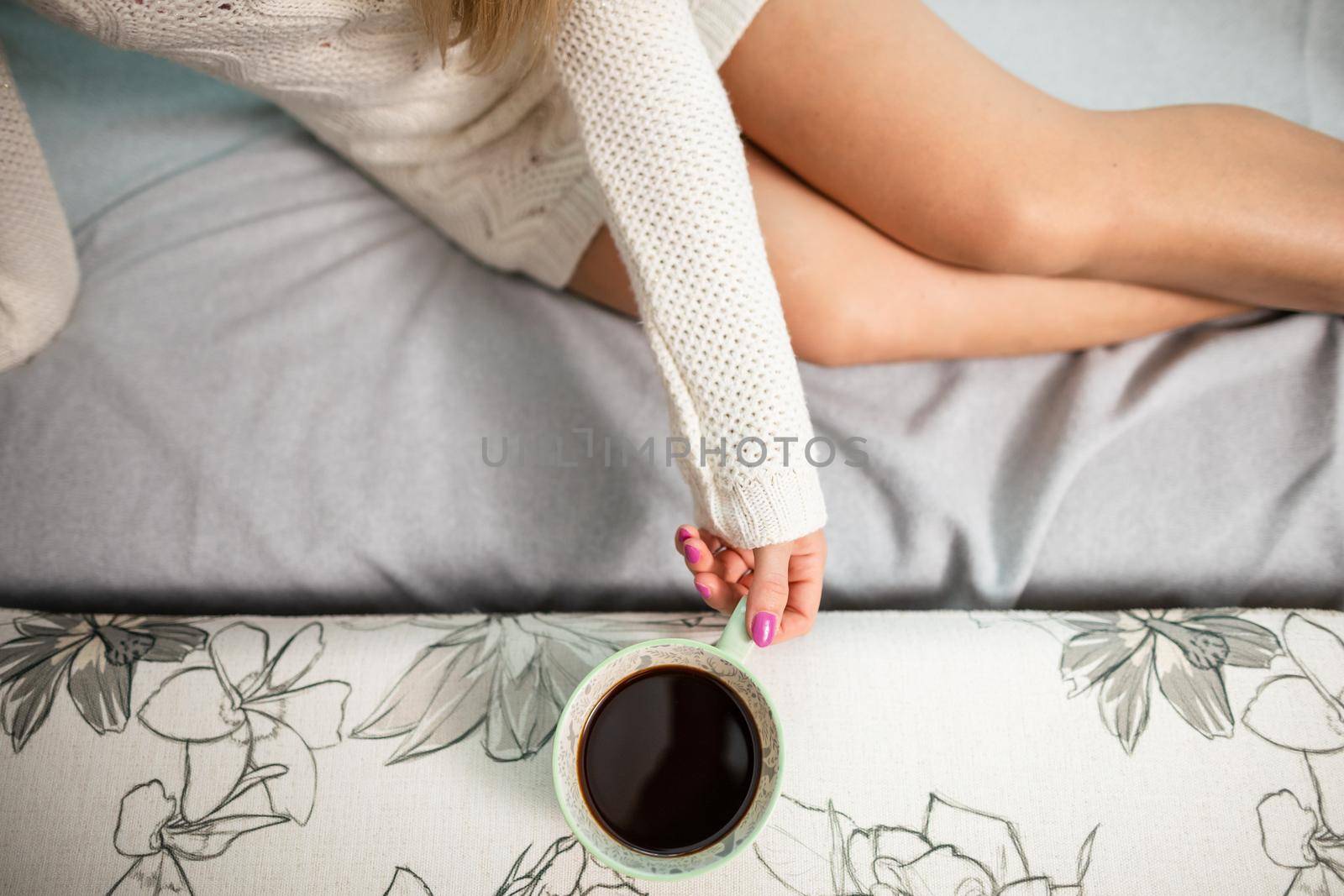 Young woman with long shapely legs is sitting on sofa at home. Porcelain cup with aromatic coffee held by woman's hand.