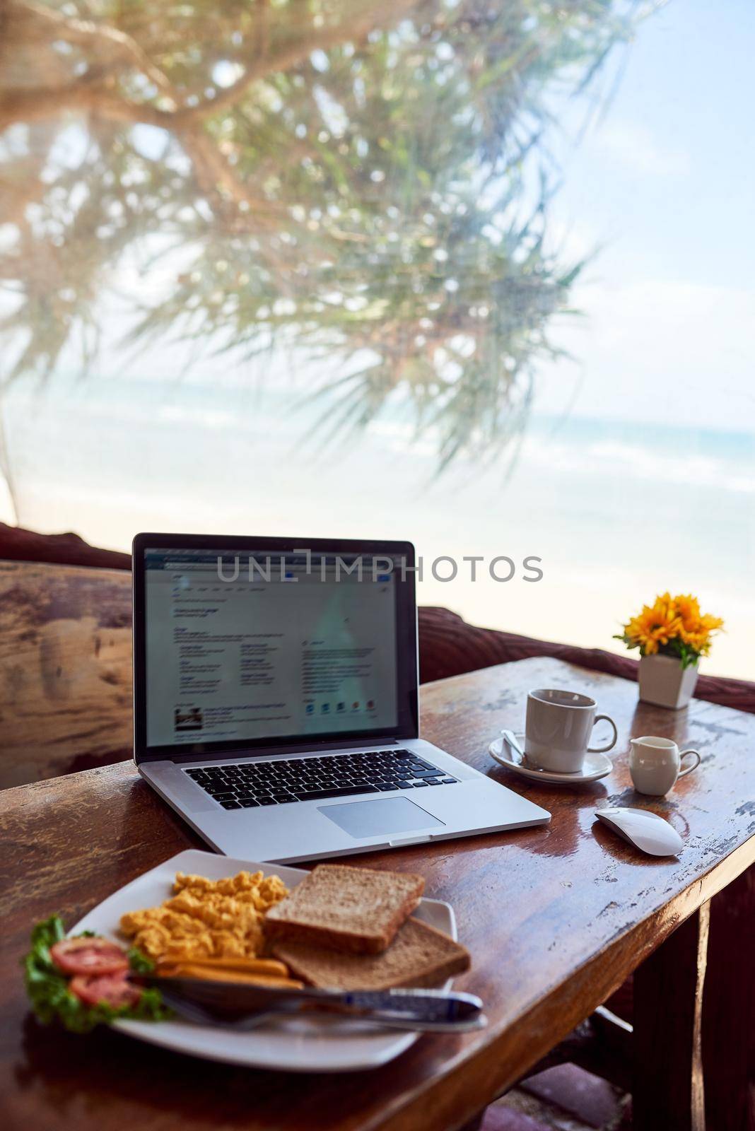 Shot of a laptop and freshly made breakfast on a table with a view of the beach in the background.