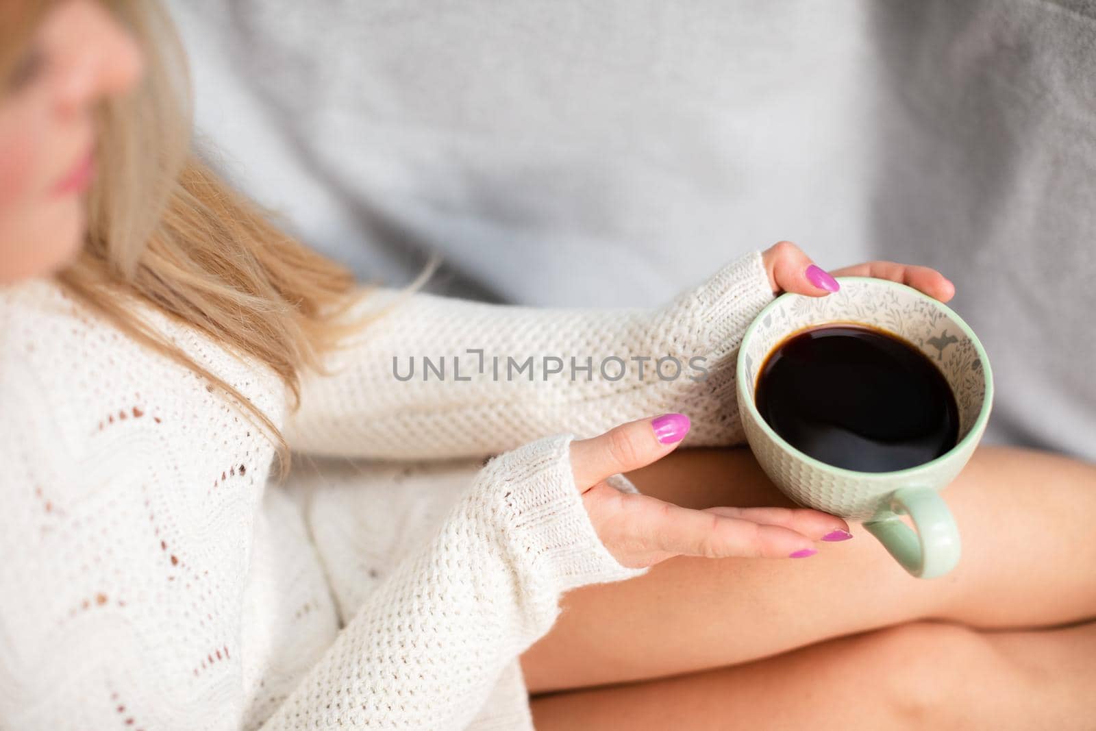 A woman with blonde hair sits on a sofa and holds a cup of coffee in her hands. Neat legs with light complexion of skin. Love for coffee and relaxation.