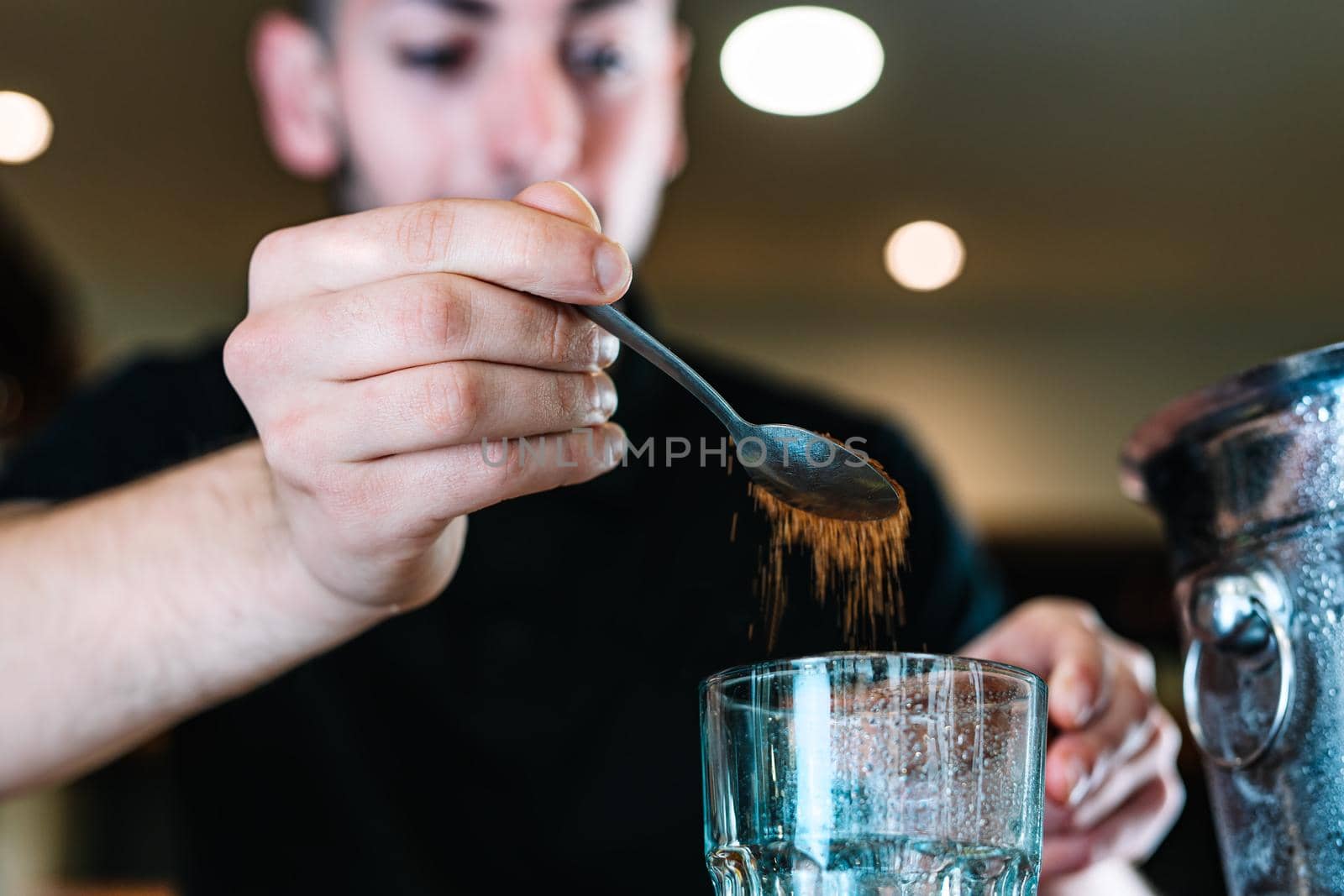 Young and modern waiter, with long dark hair, dressed in black polo shirt, adding brown sugar in a large crystal glass to prepare a cocktail. Waiter preparing a cocktail. Cocktail glass with ice cubes. Mojito. Bar full of cocktail ingredients. Dark background and dramatic lighting. Horizonta