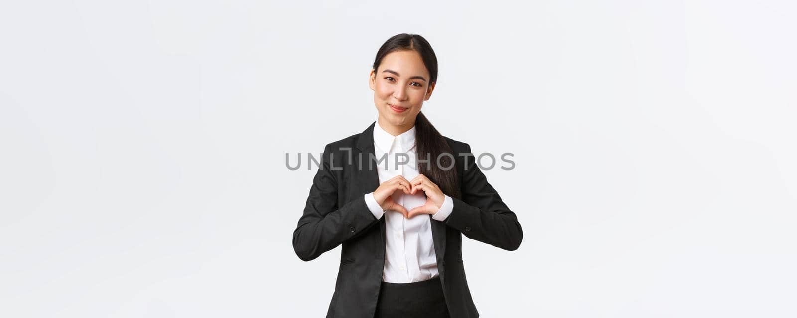 Close-up of pleasant asian businesswoman, saleswoman in black suit care for her clients, showing heart sign and smiling with admiration, standing white background caring and tender by Benzoix