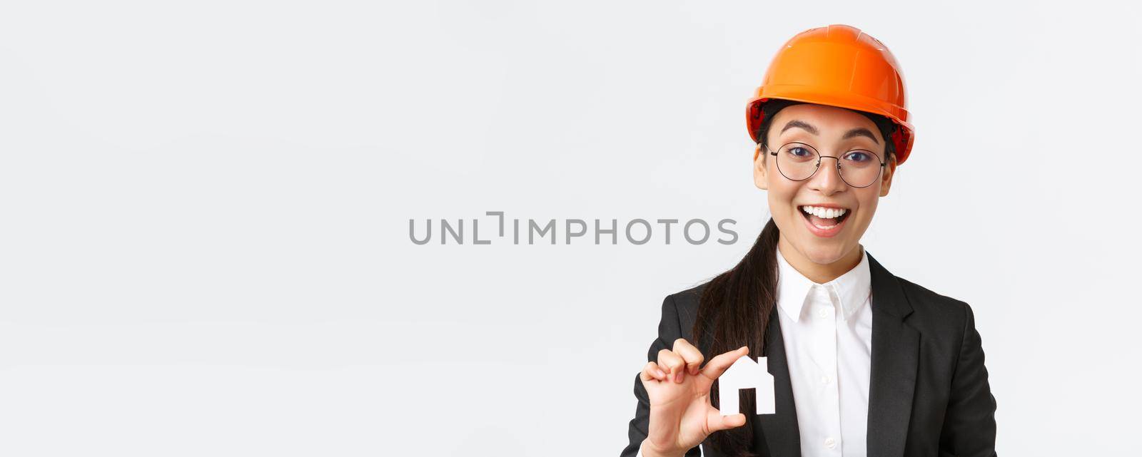 Close-up of professional broker in safety helmet and business suit selling homes. Cheerful architect showing minuature of house and smiling, working over new design or construction, white background.