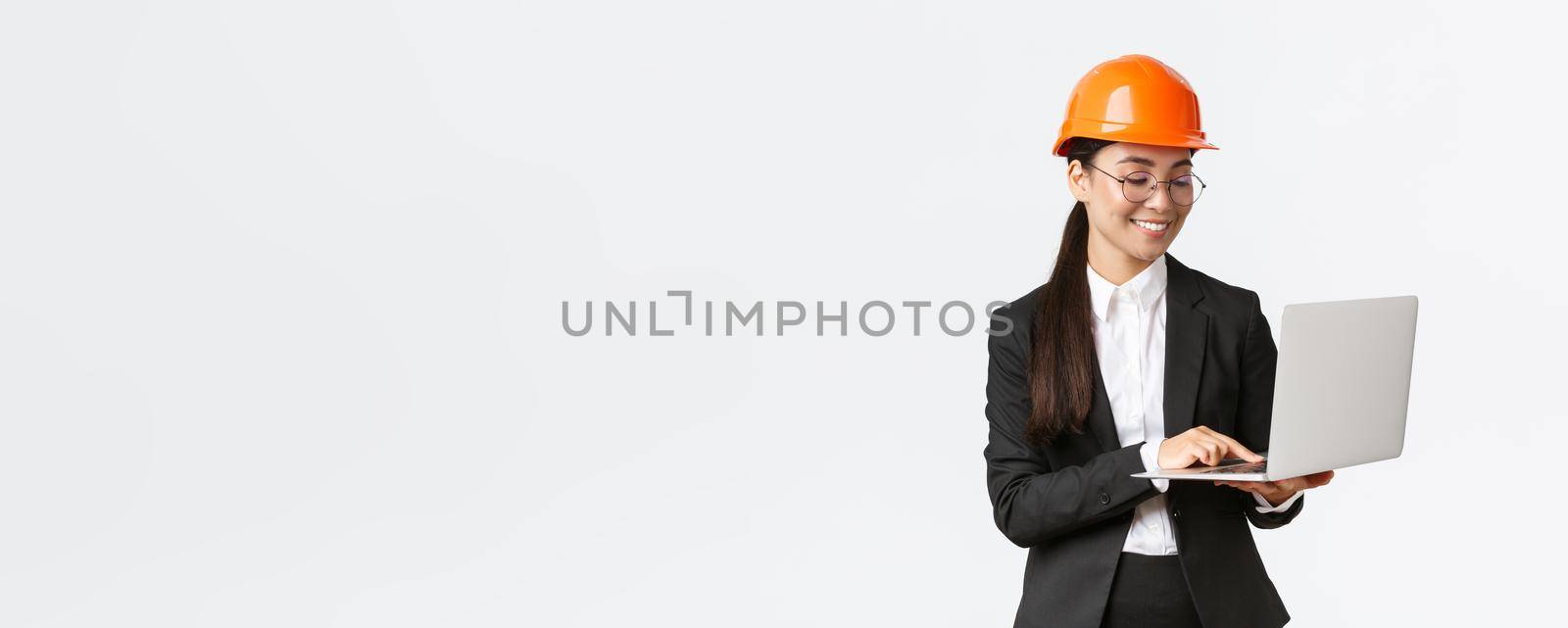 Smiling successful female asian industrial engineer, factory manager in safety helmet and business suit using laptop computer, checking with project or blueprints on screen by Benzoix