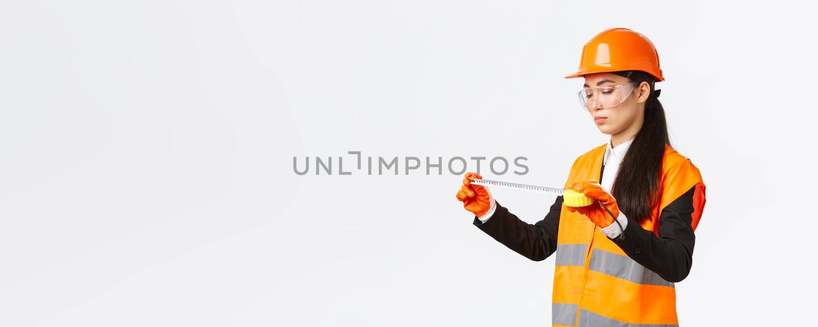 Serious asian female construction engineer, technician inspect layout, measuring something, looking at tape measure with focused face, standing over white background in safety uniform.