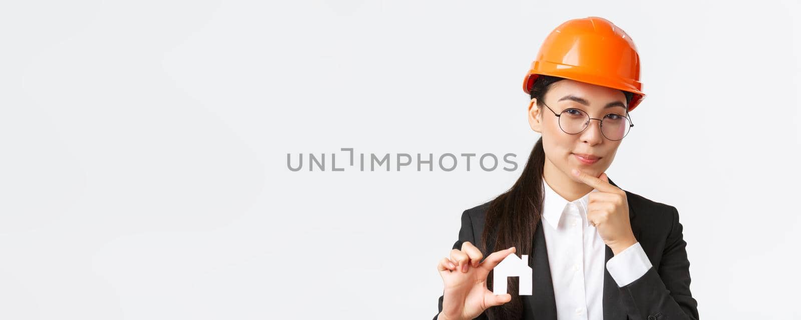 Thoughtful creative female asian architect in helmet and business suit creating new ideas of design, showing home maket and thinking, making cunning face, pondering plan of construction by Benzoix