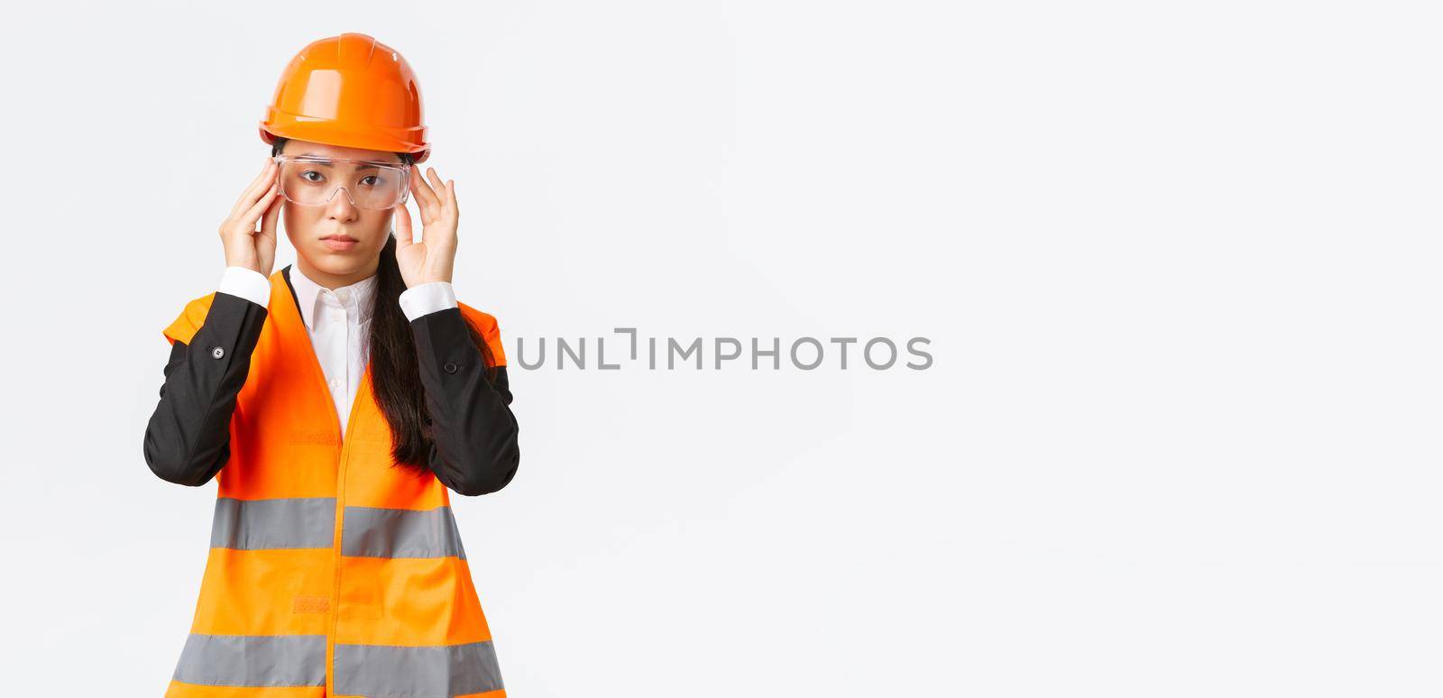 Serious-looking young asian female construction manager follow safety protocol, wearing protective glasses and helmet before entering enterprise, standing white background.