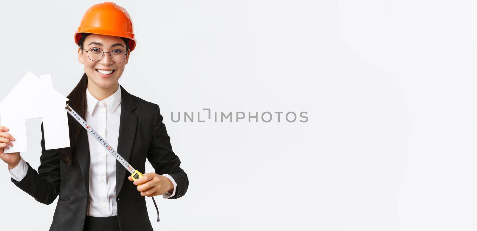 Smiling professional female asian engineer, architect in helmet and business suit showing house maket and tape measure, ready starting home renovation, construction works, white background.