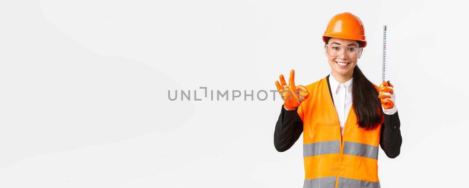 Satisfied asian female construction engineer, architect or inspector at enterprise showing okay gesture and tape measure, smiling pleased, give permission, approve measurements are valid by Benzoix
