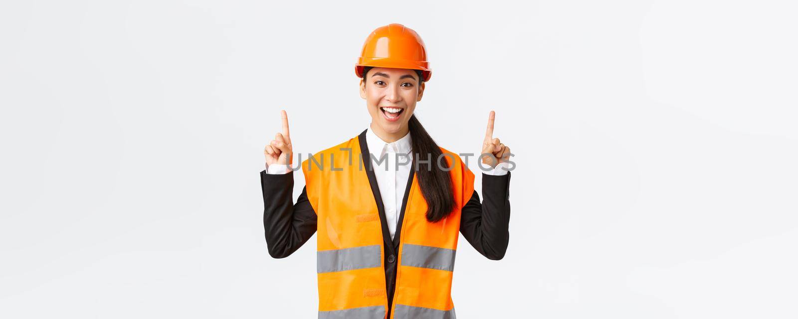 Building, construction and industrial concept. Happy smiling female asian engineer in safety helmet and reflective clothing, introduce new object, estate for sale. Architect pointing fingers up.