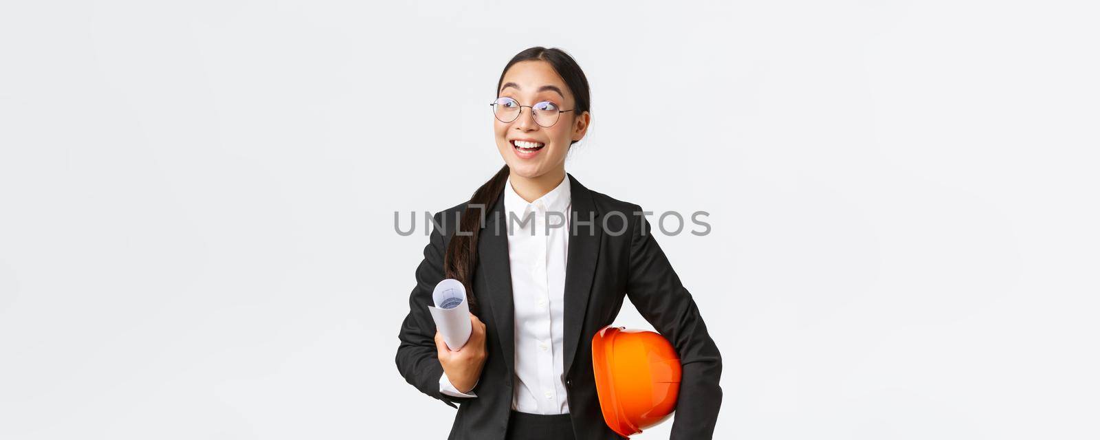 Hopeful and excited asian female architect looking around with fascination and amazement, holding blueprints and helmet, walking in construction area, standing in business suit over white background.